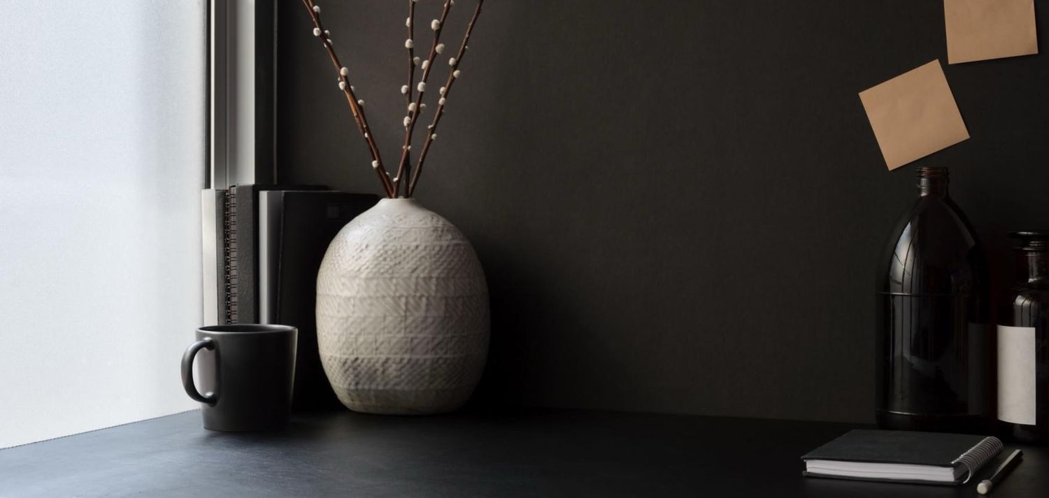 Minimalist dark desk with textured ceramic vase, black mug, decorative bottle, and dried branches against a dark wall.