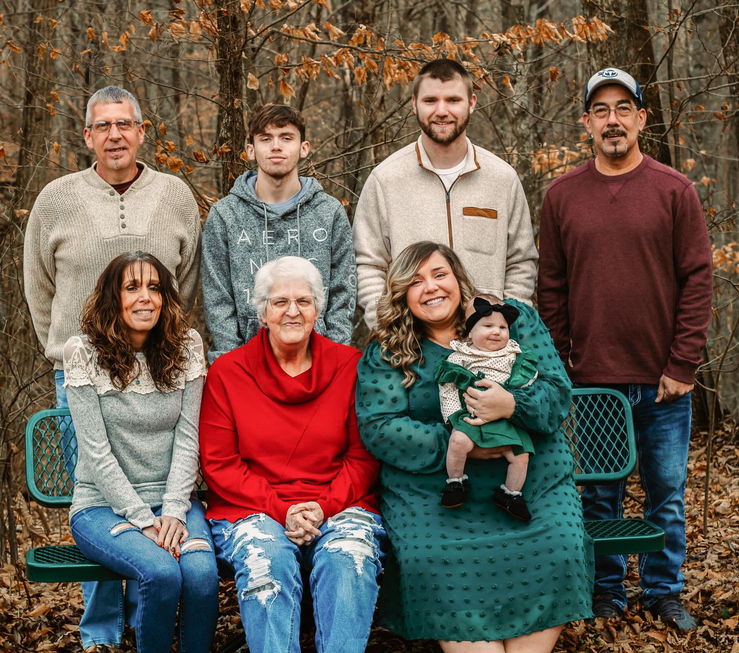 Multi-generational family portrait outdoors in autumn woods, seven members posing together, including baby in green outfit.