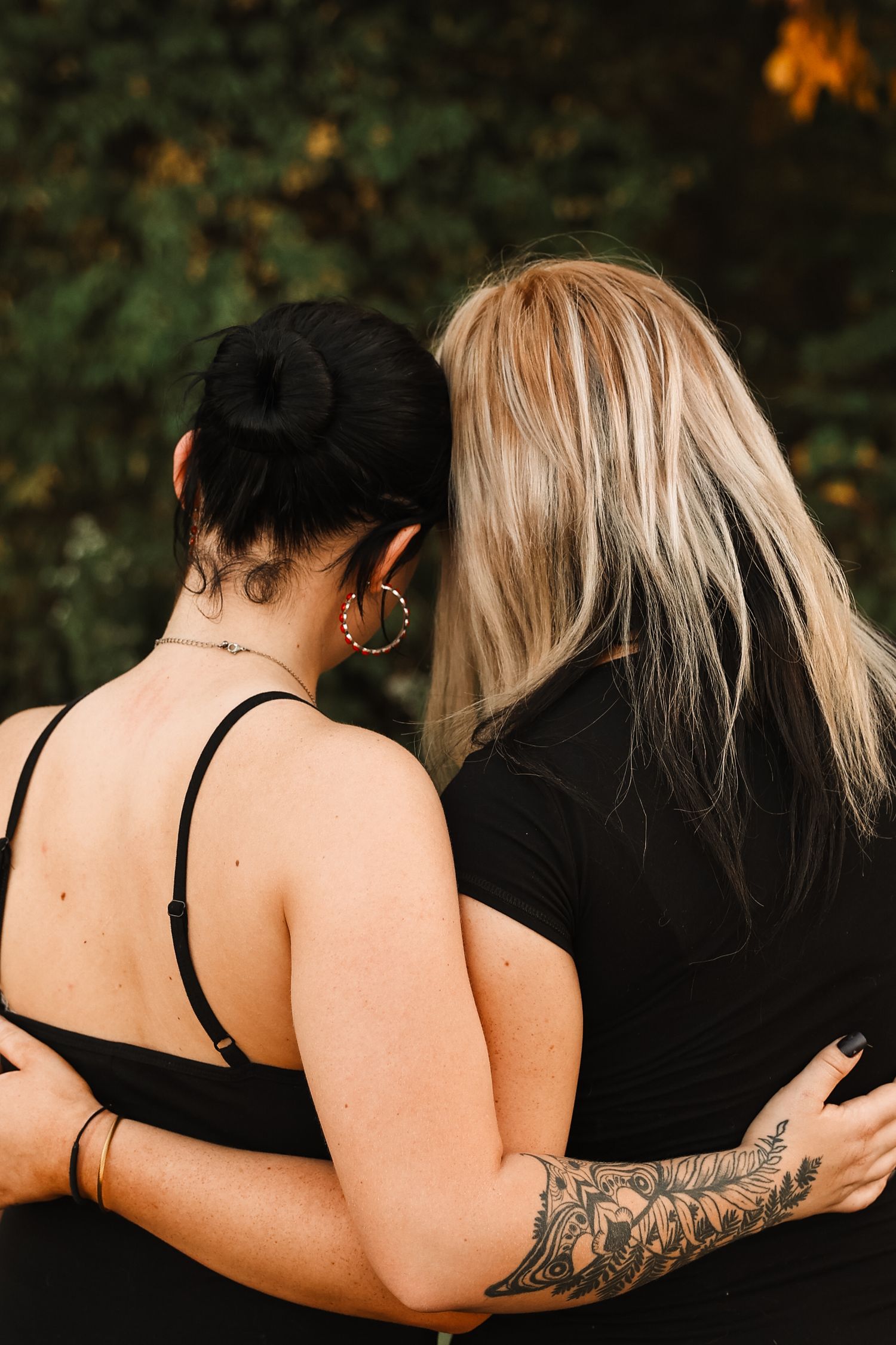 Two women with black and blonde hair embrace outdoors, one with a tattoo sleeve, both wearing black tops.