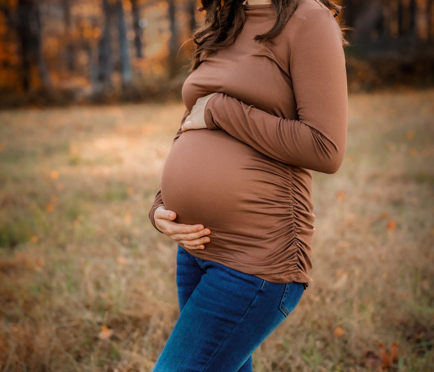 Pregnant woman in brown top and jeans cradling baby bump outdoors in autumn field.