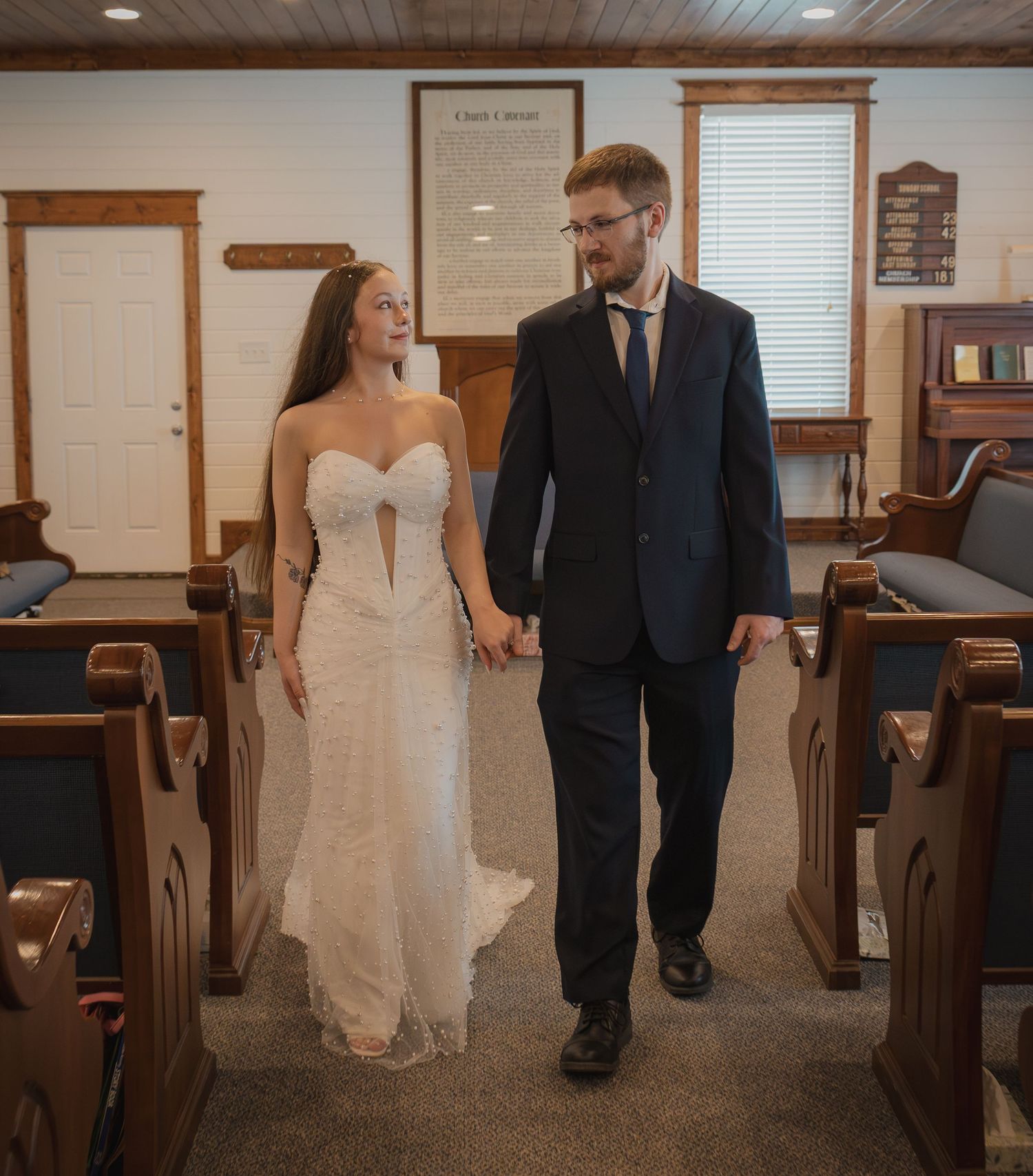 Bride and groom walk hand in hand down church aisle, she in white gown, he in navy suit.