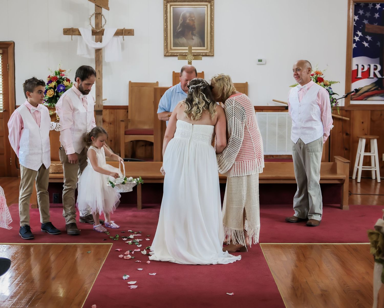 Bride in white gown stands with family at church altar during wedding ceremony, flower girl nearby on red carpet aisle.