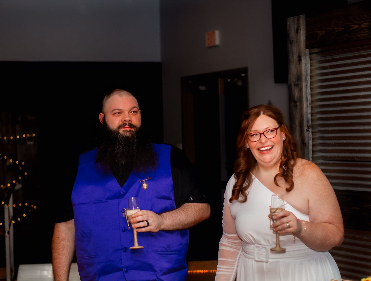 Smiling couple at a wedding reception, man in blue vest with beard holding champagne, woman in white dress with glasses.