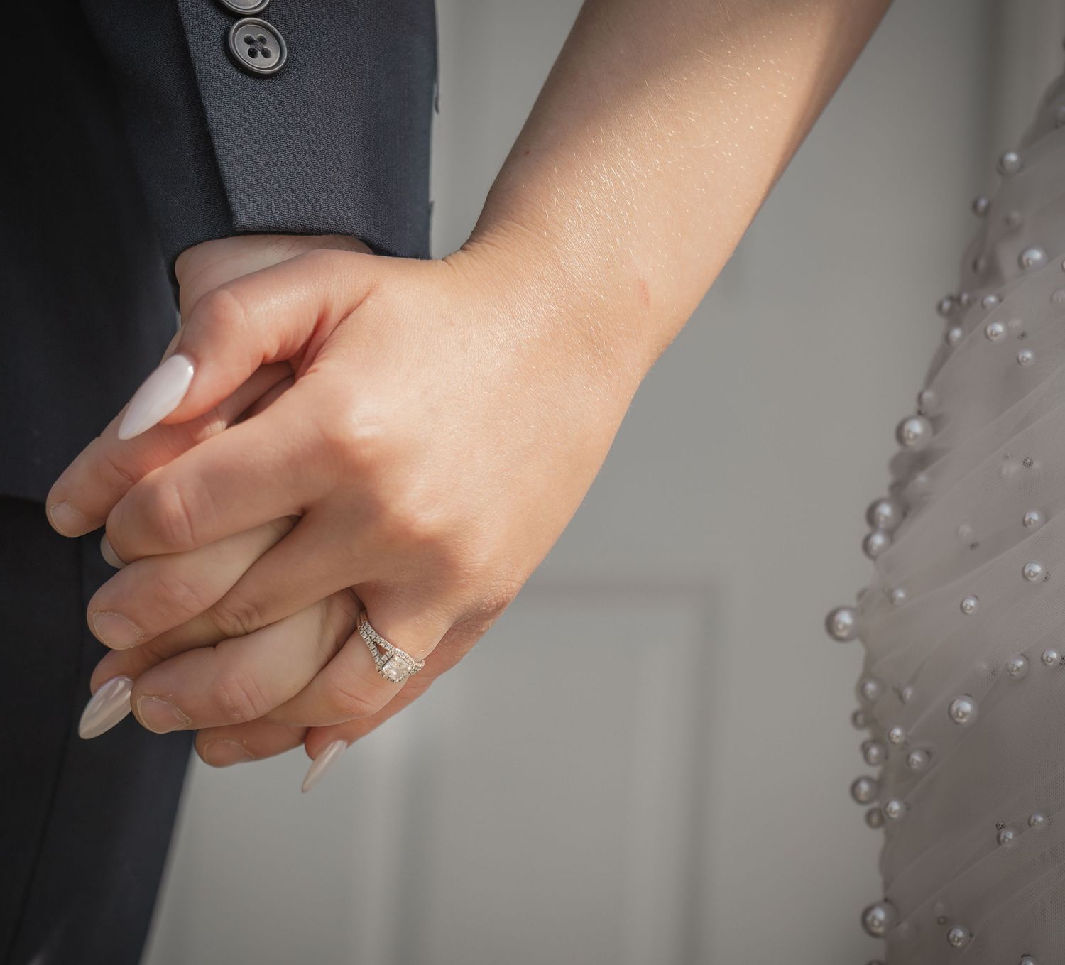 Close-up of a couple holding hands, woman wearing a diamond engagement ring, man in a gray suit, bride in a pearl-adorned dress.