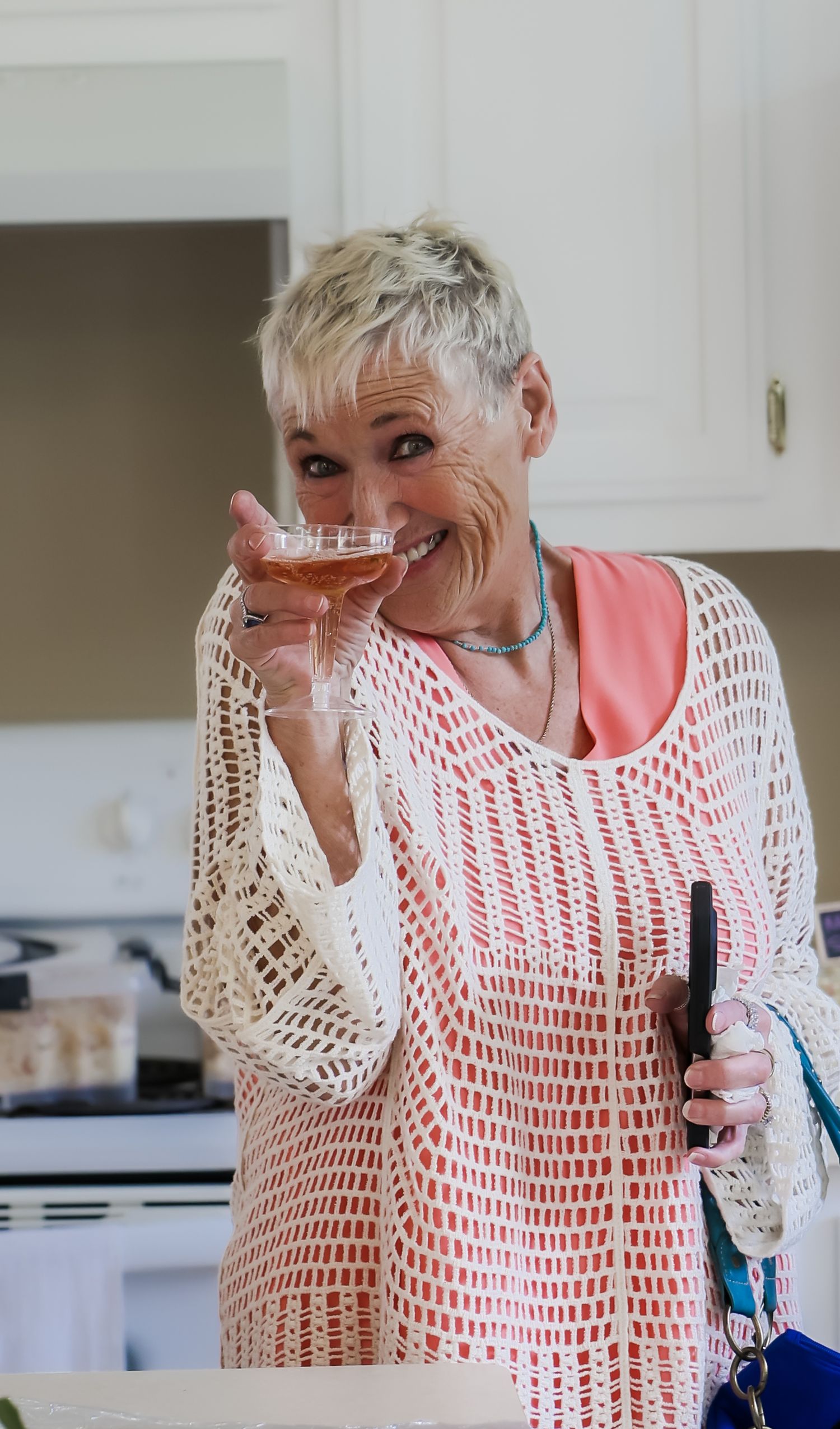 Smiling older woman with short blonde hair holding a wine glass in a bright kitchen wearing a white crochet top.