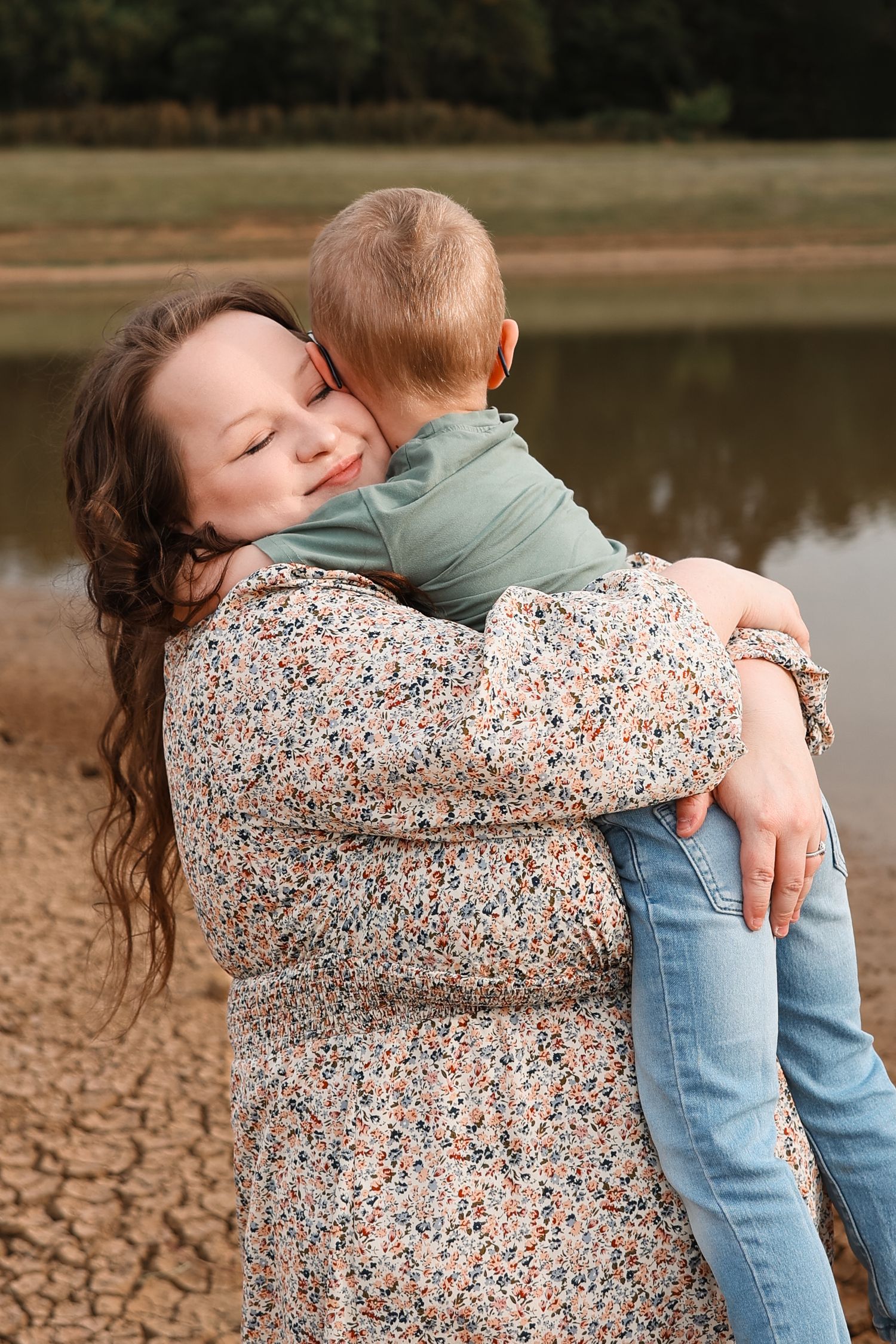 A mother with closed eyes joyfully hugs her young child near a lake, both wearing casual floral and denim outfits.