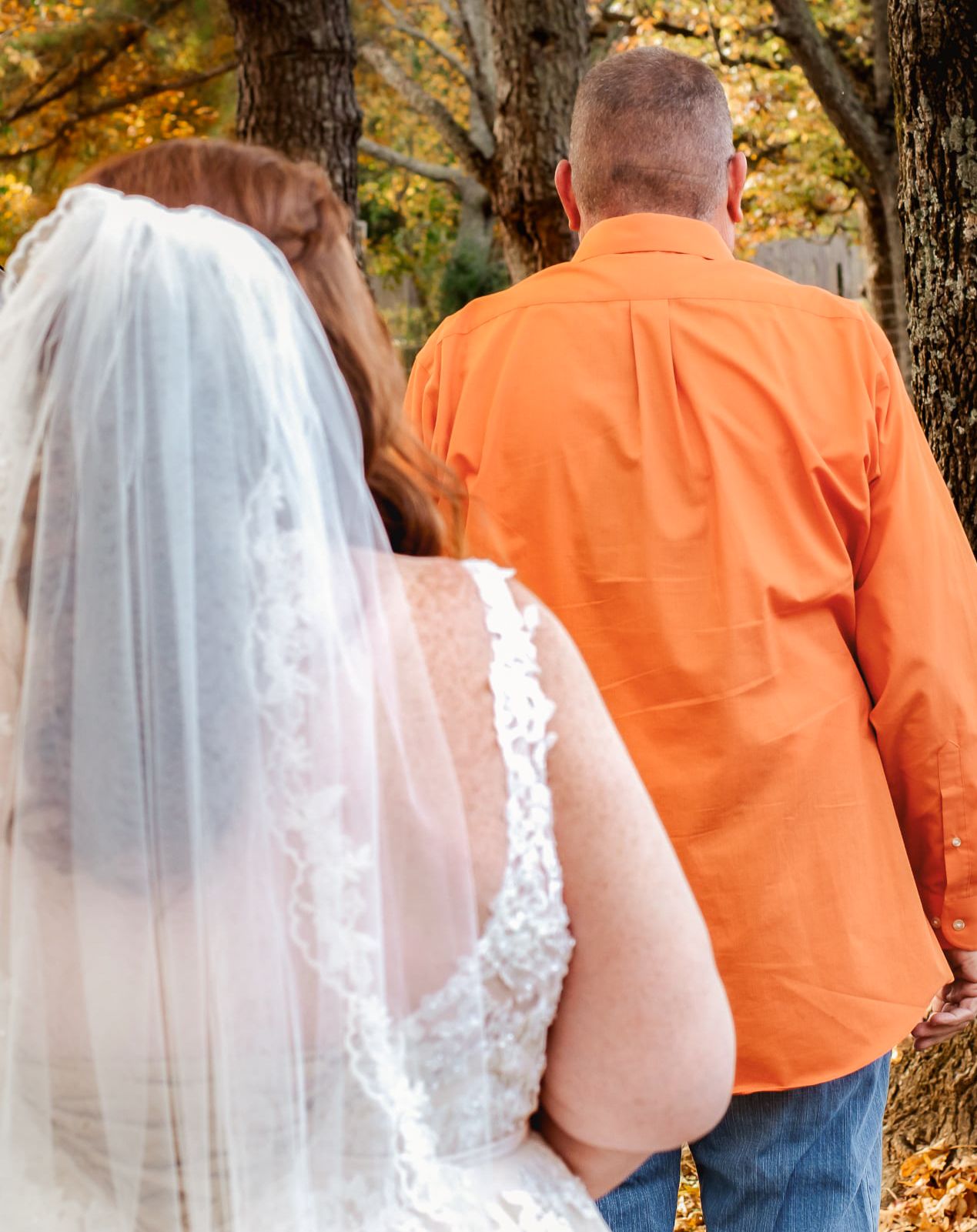 Bride in white lace gown and veil stands beside groom in orange shirt among autumn trees.