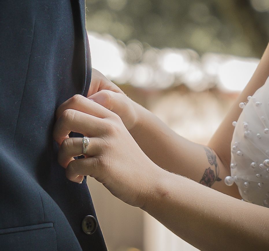 Bride with diamond ring and floral tattoo adjusts groom's navy suit jacket at outdoor wedding ceremony.