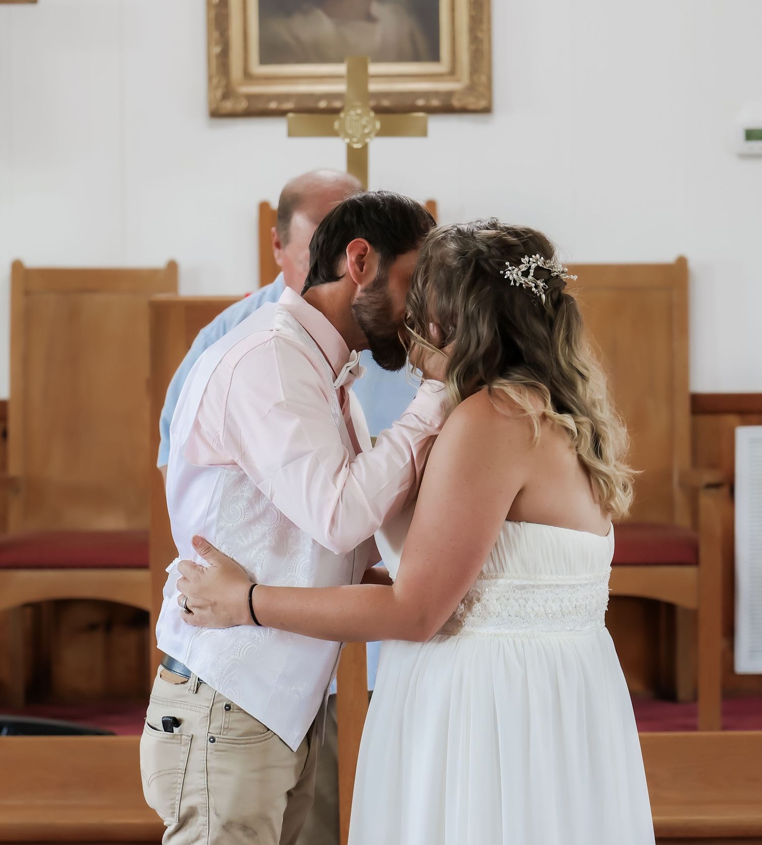 Couple sharing first kiss at church wedding altar, groom in pink shirt, bride in white dress with floral hairpiece.