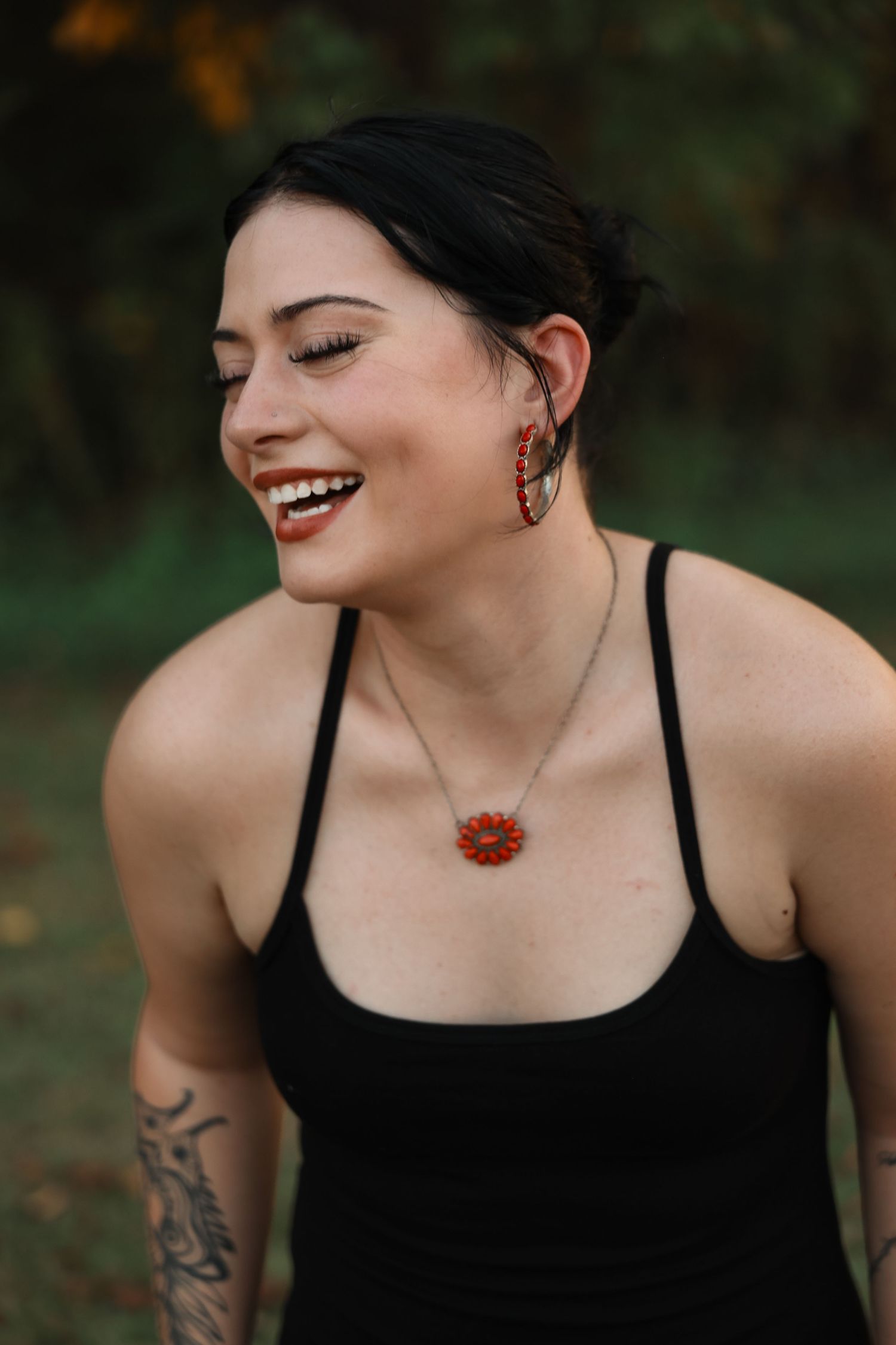Smiling woman with black hair wearing red floral earrings, matching necklace, black tank top, and arm tattoo outdoors.