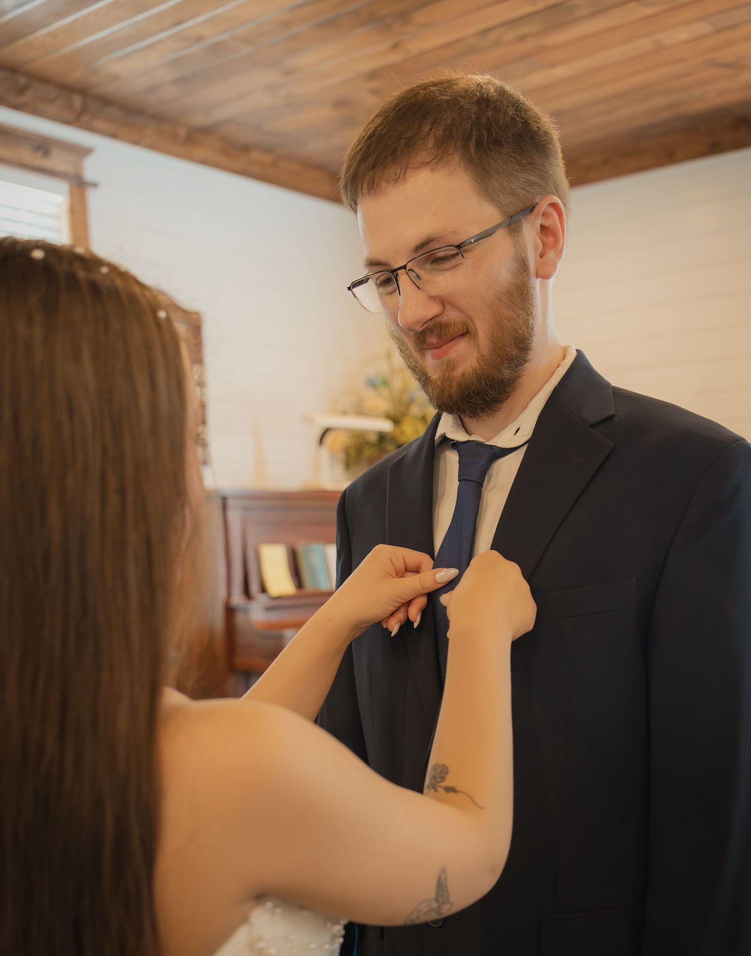 Bride adjusting groom's blue tie on wedding day, man in navy suit smiling, rustic indoor venue background.