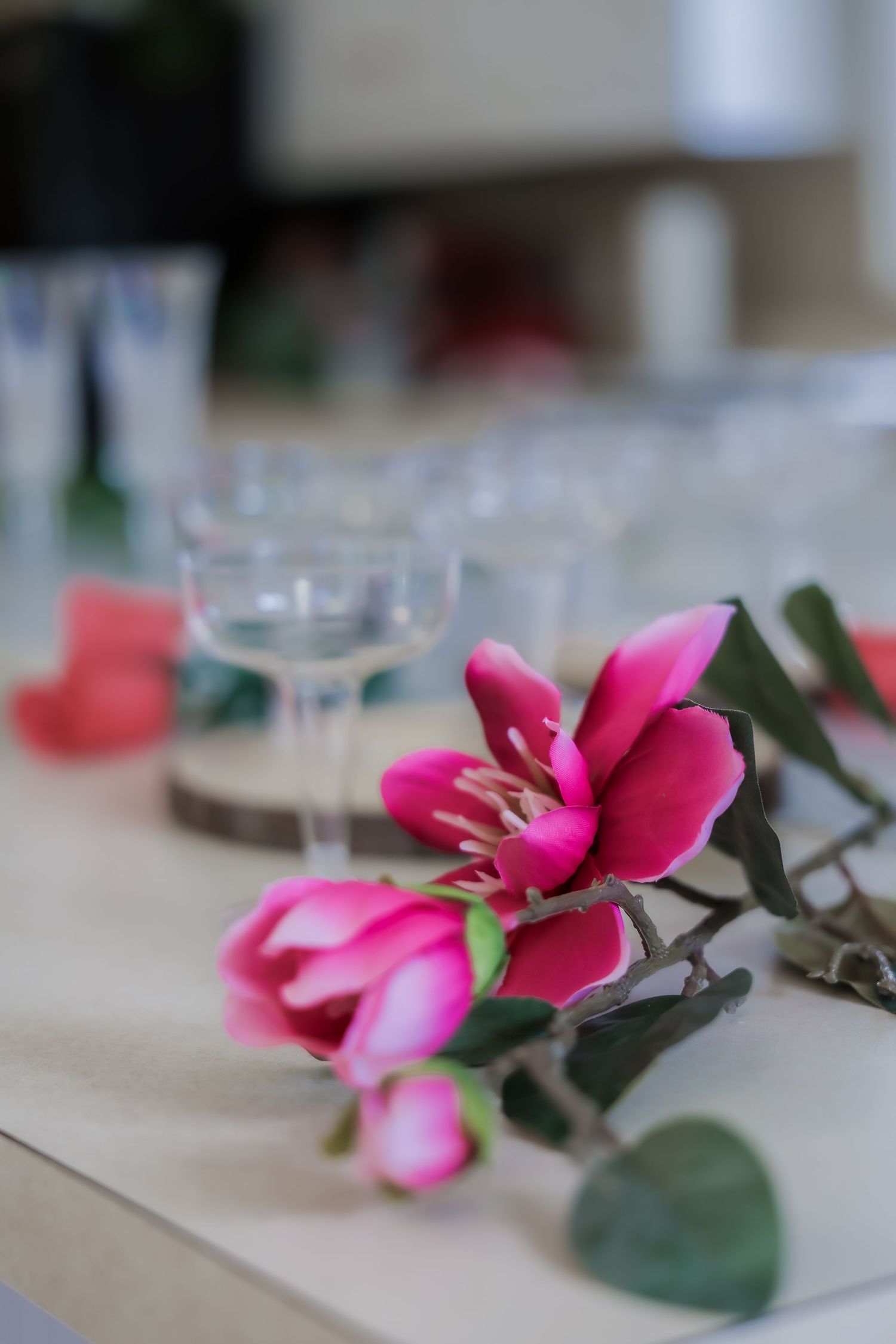Pink artificial magnolia flowers beside clear glass candle holders on a white table.