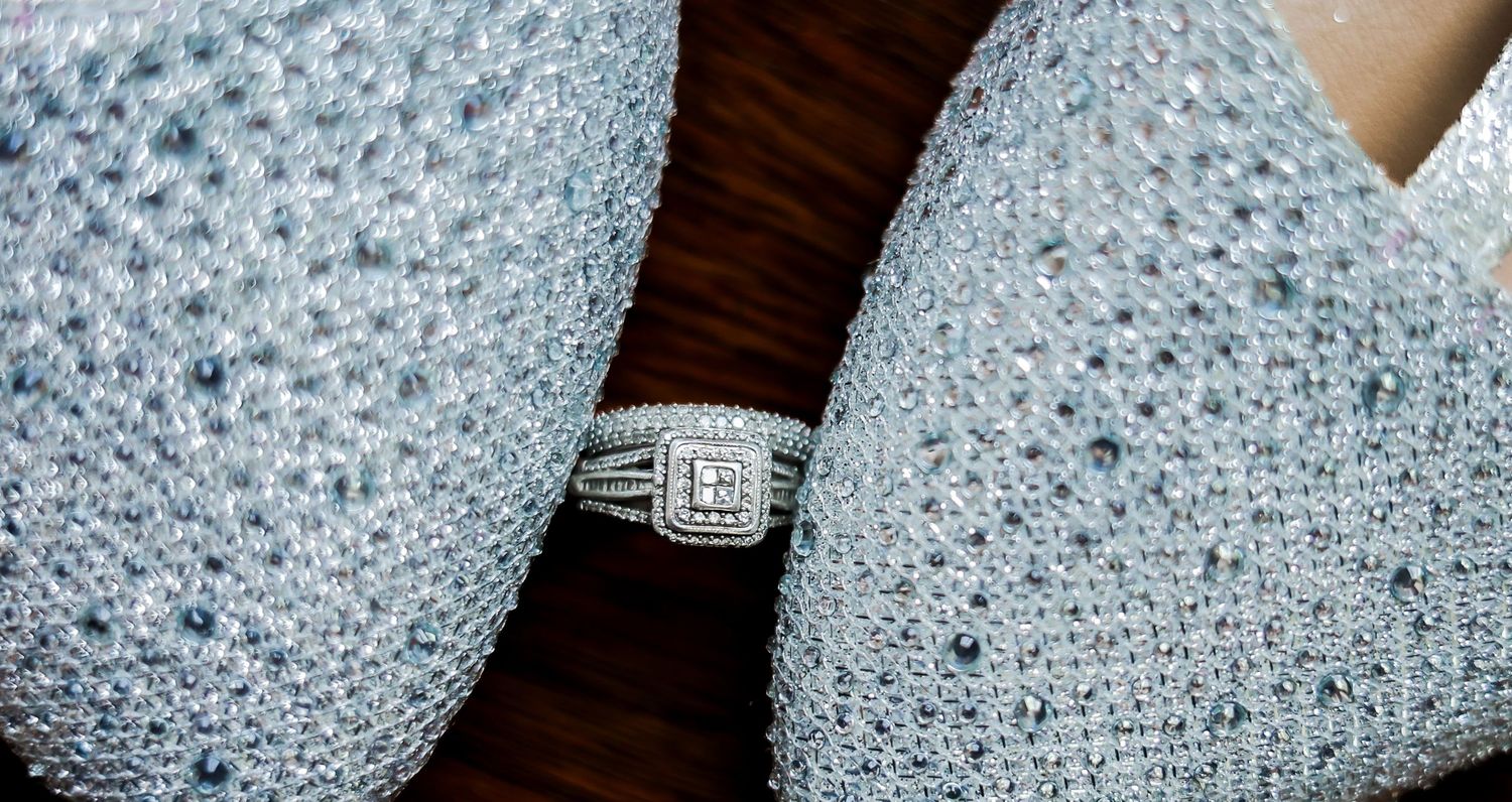 Diamond halo engagement ring held between two sparkling rhinestone-covered gloves on a wooden background.