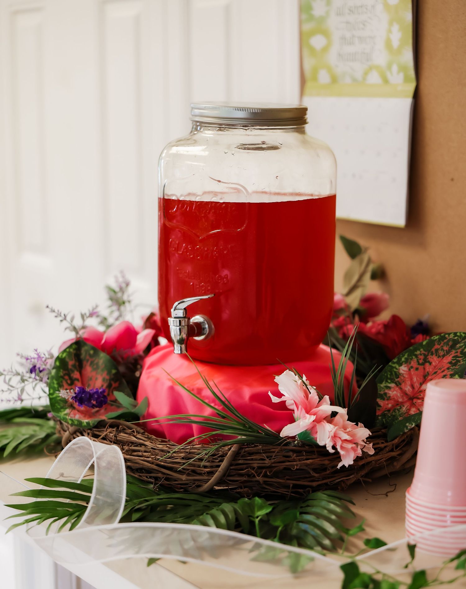 Glass drink dispenser filled with red punch, surrounded by a floral wreath centerpiece with pink flowers and greenery.