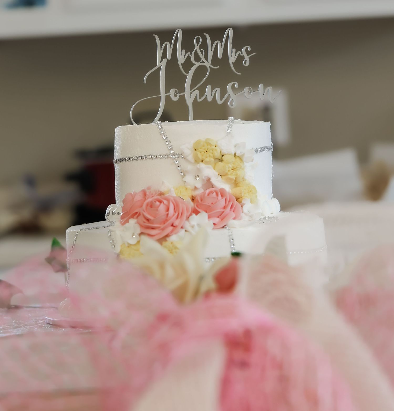 White two-tier wedding cake with pink roses and Mr & Mrs Johnson cake topper, decorated with floral accents and pink tulle.