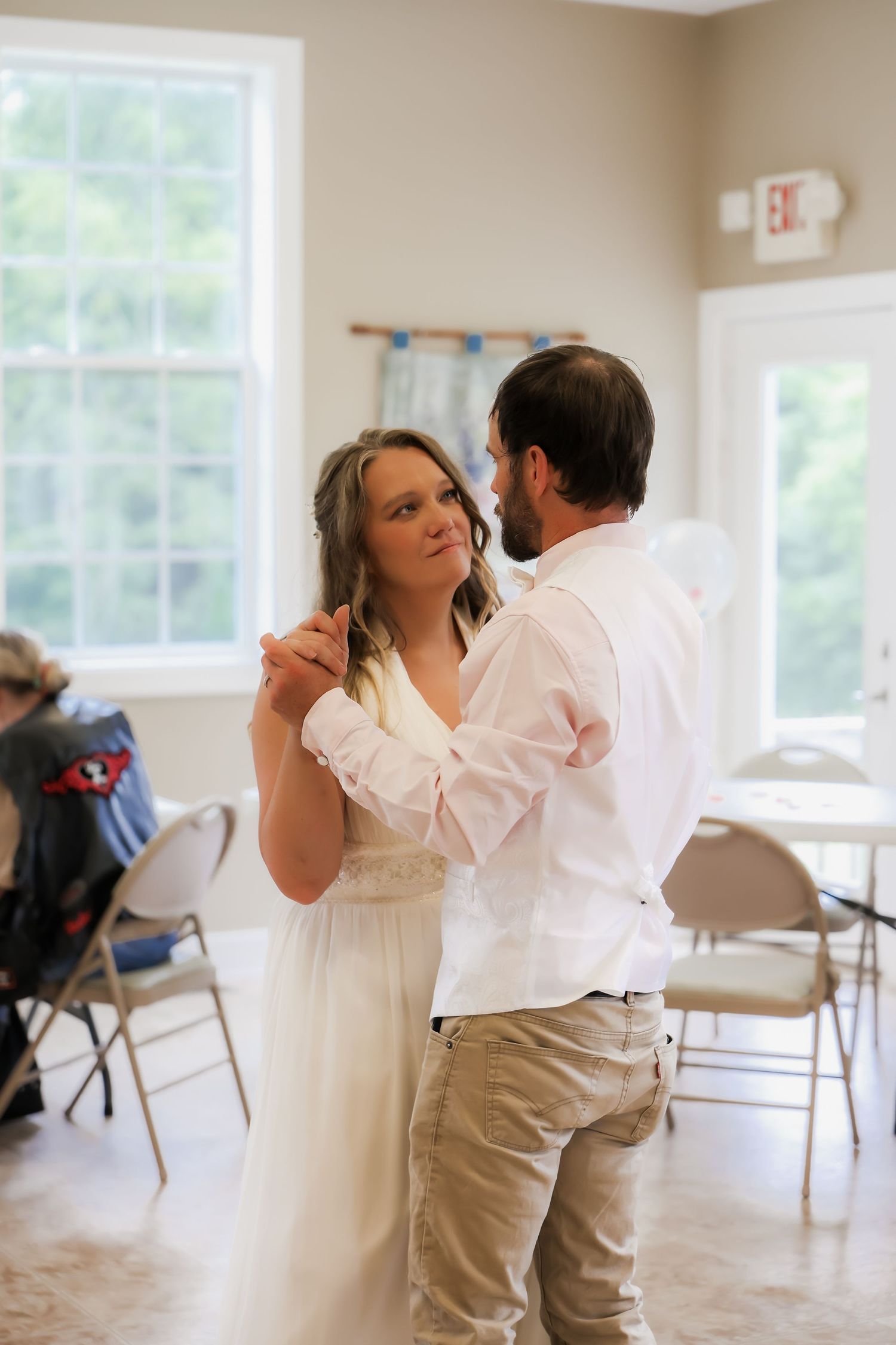 Bride and groom share their first dance in a bright reception hall, dressed in white and khaki attire.