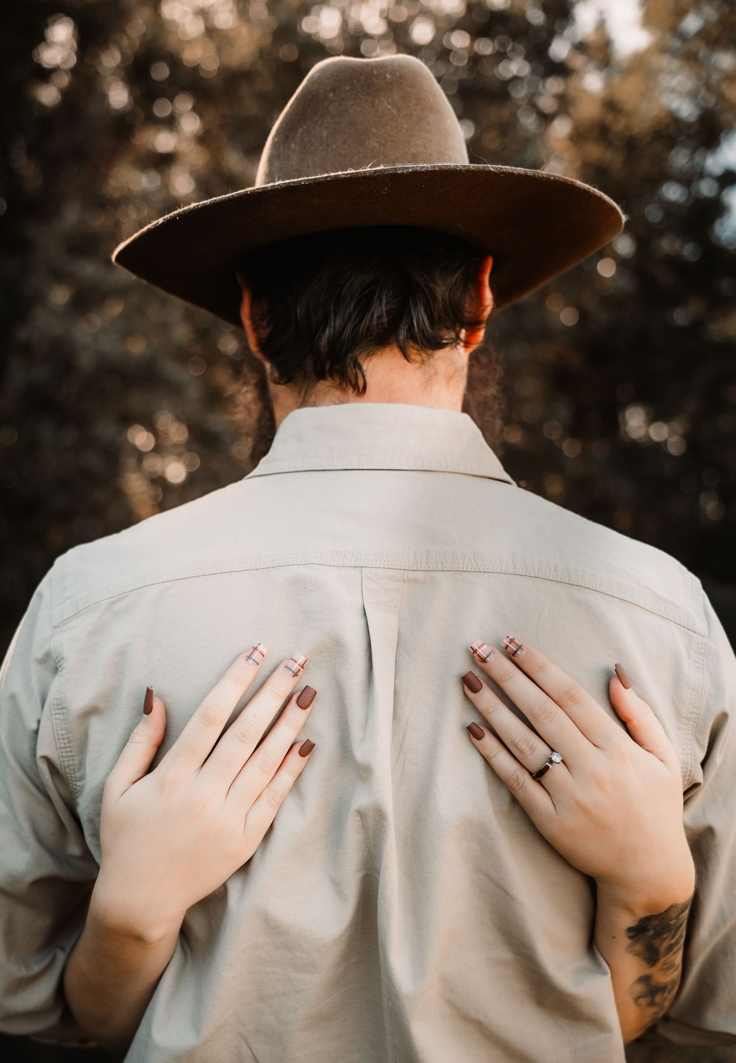 A woman places her hands on a man's back as he faces away wearing a wide-brim hat and light button-up shirt.