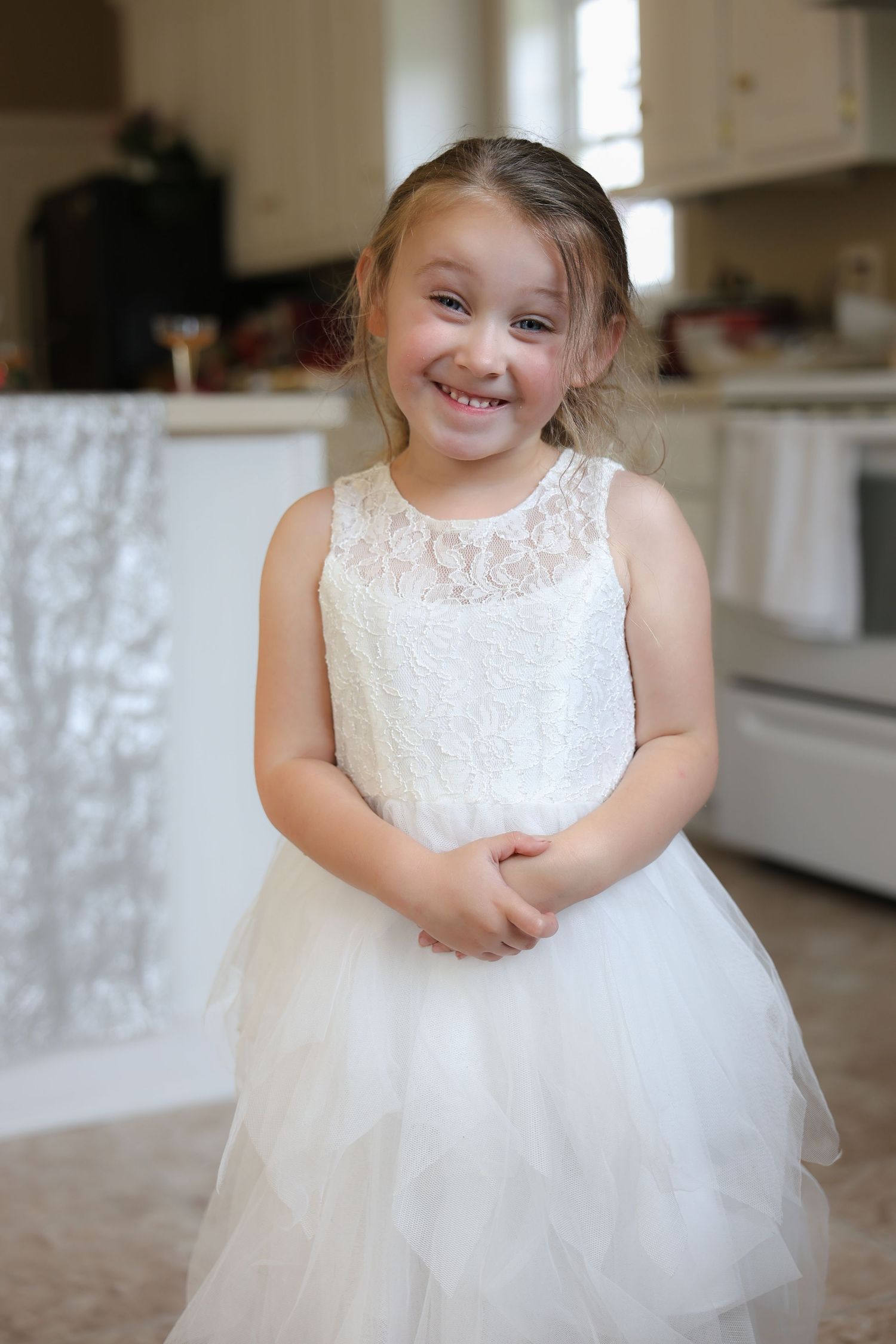 A smiling young girl in a white lace and tulle flower girl dress poses happily in a bright kitchen.