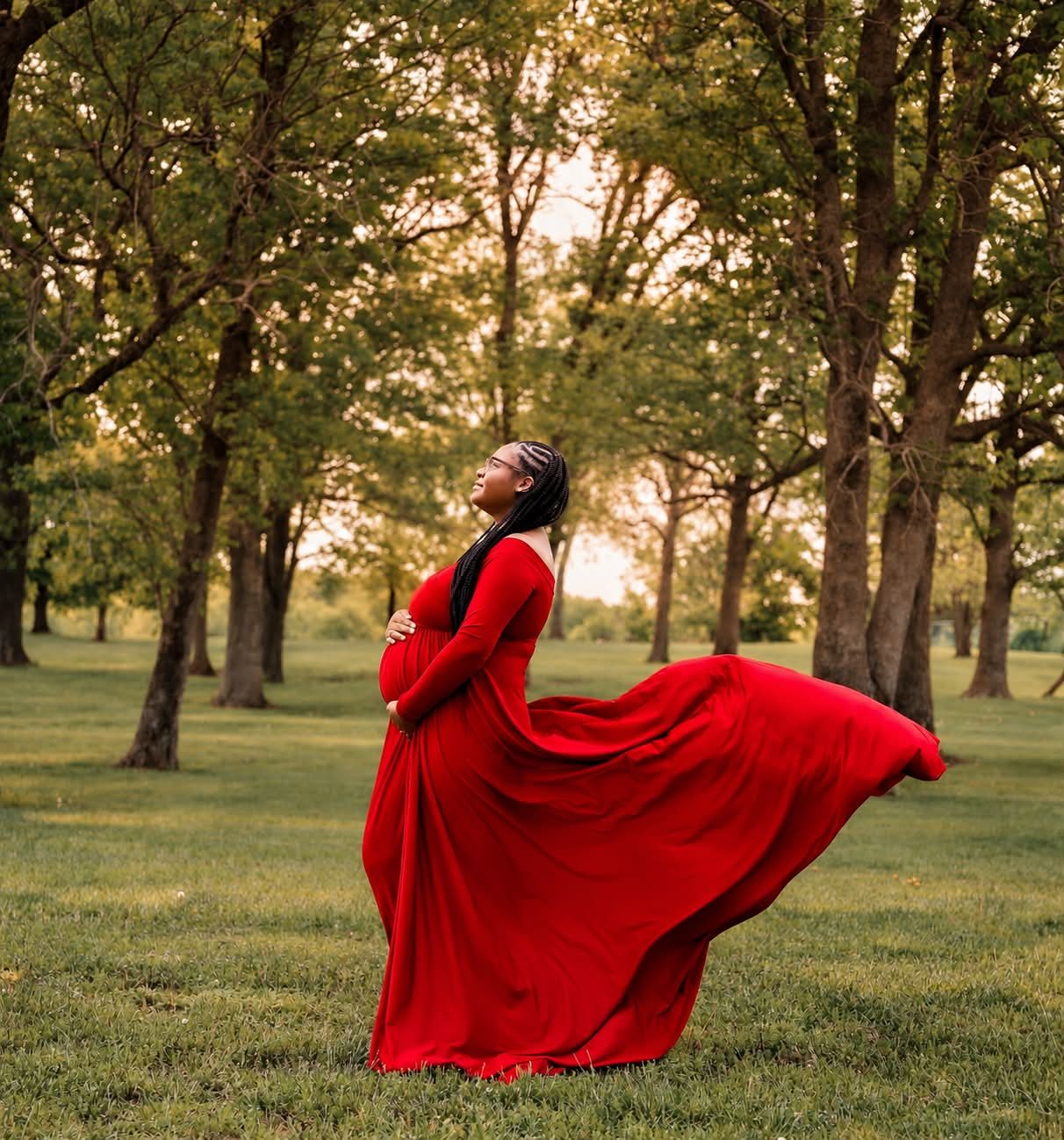 Pregnant woman in flowing red gown twirling in lush green park surrounded by trees during golden hour.