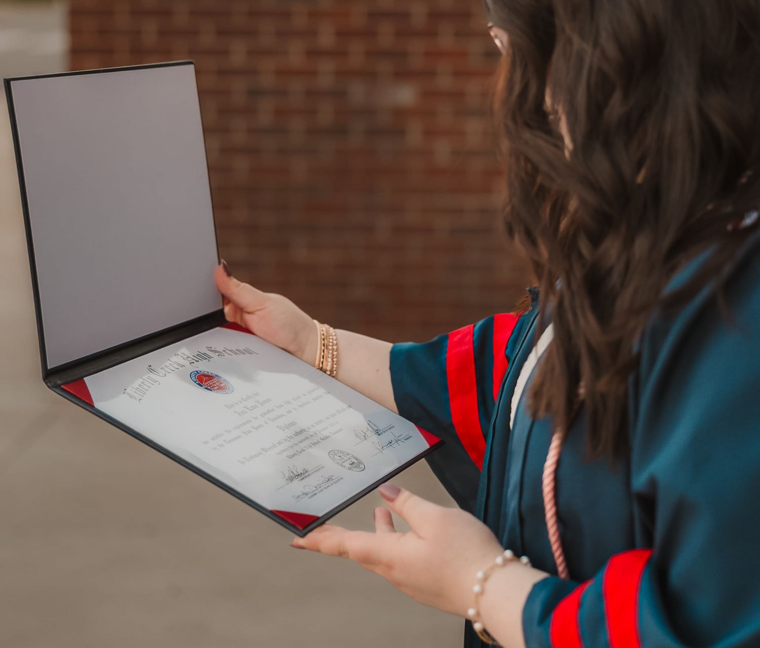 Young woman in teal jacket holding open a diploma in a black folder outdoors near a brick wall.