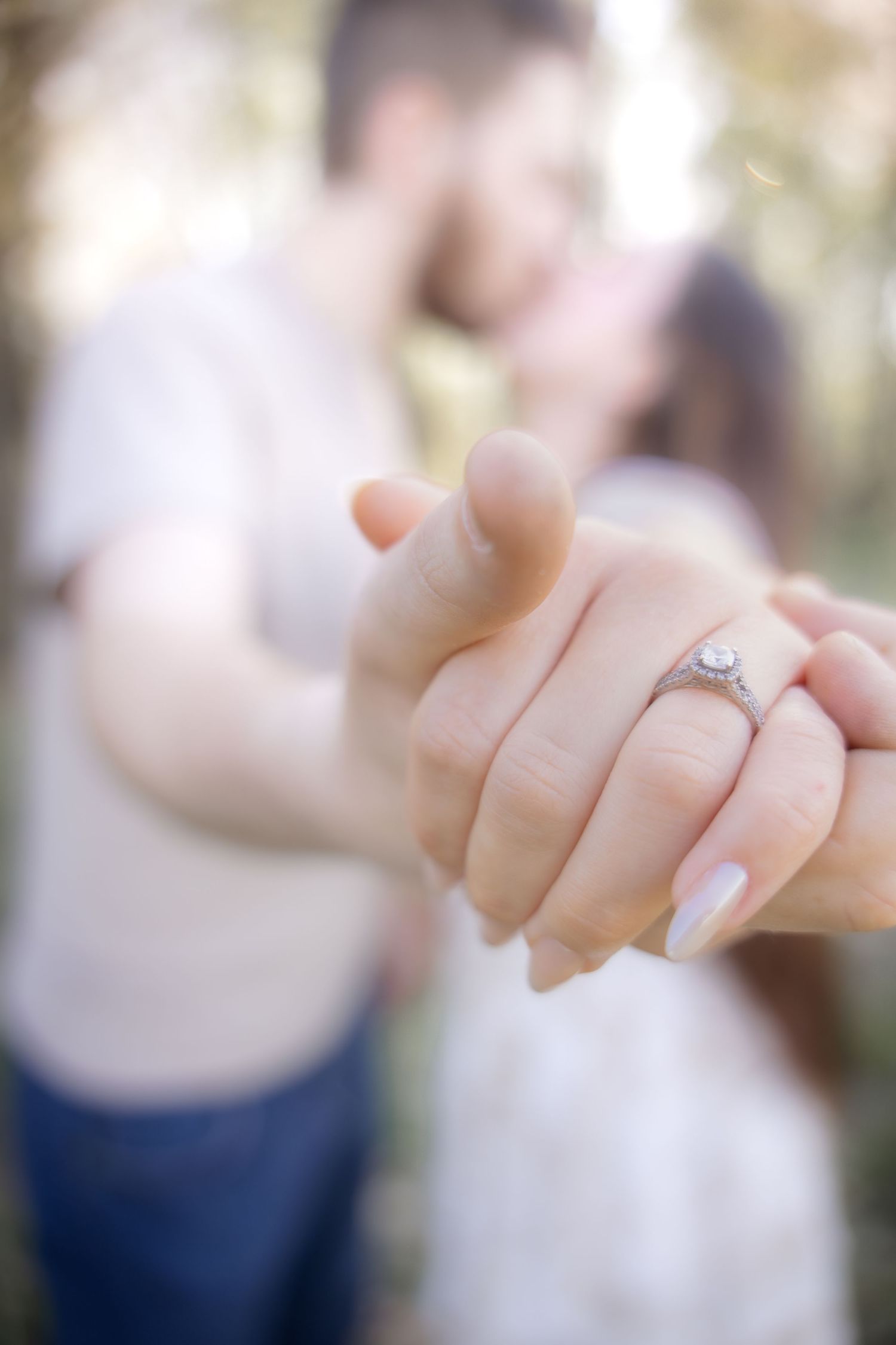 Close-up of an engagement ring on a woman's hand while a couple kisses softly in the blurred background.