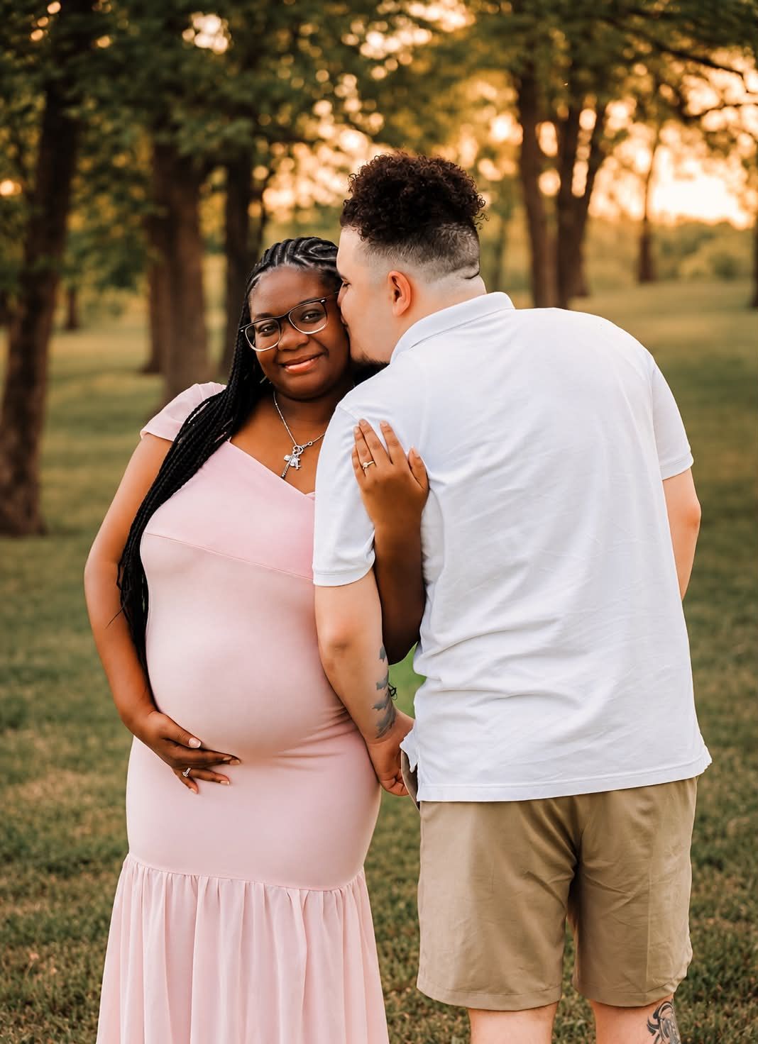 A pregnant woman in a pink dress smiles as her partner kisses her cheek during a golden hour maternity photo shoot.