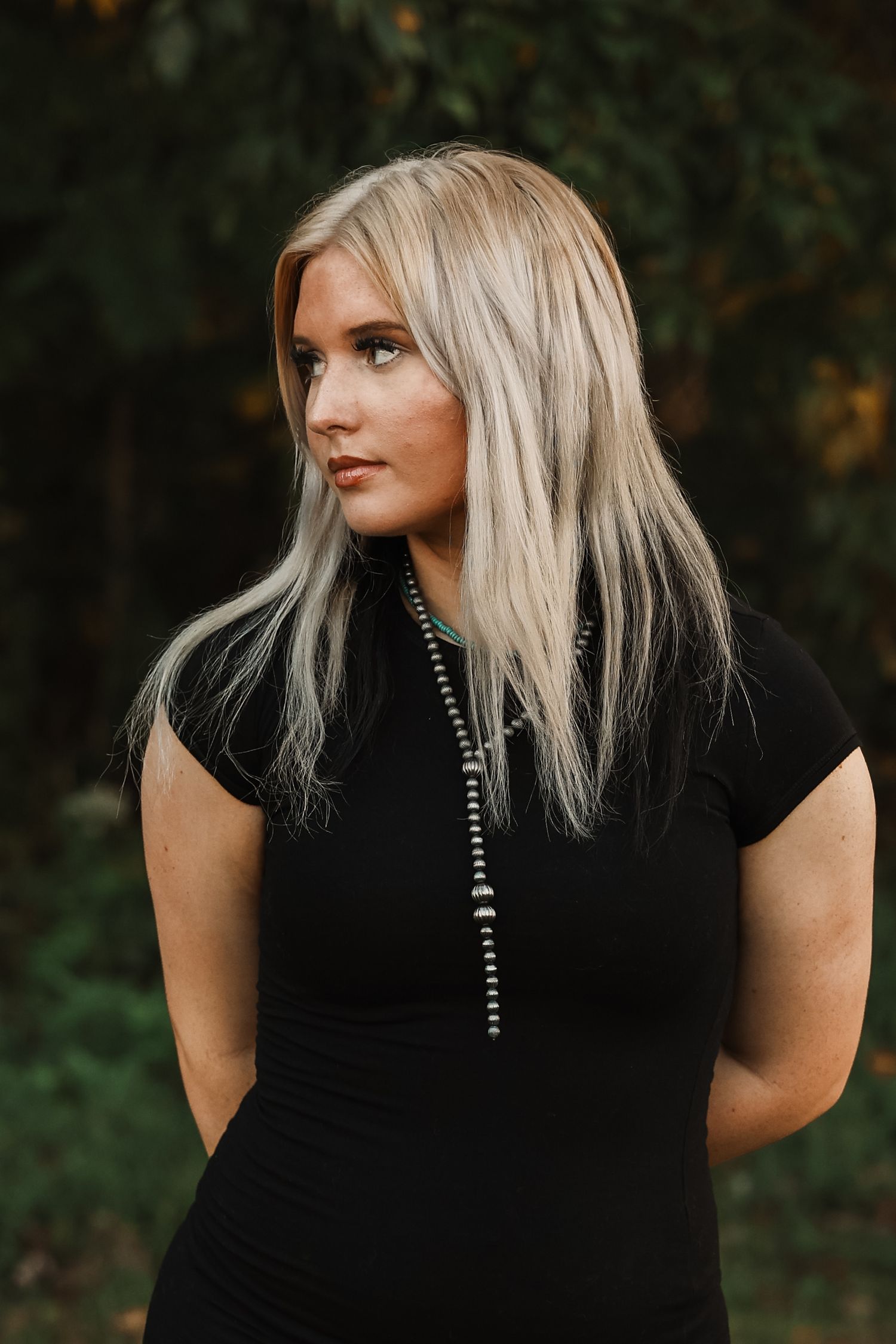 Blonde woman in black dress with beaded necklace posing outdoors against green foliage background.