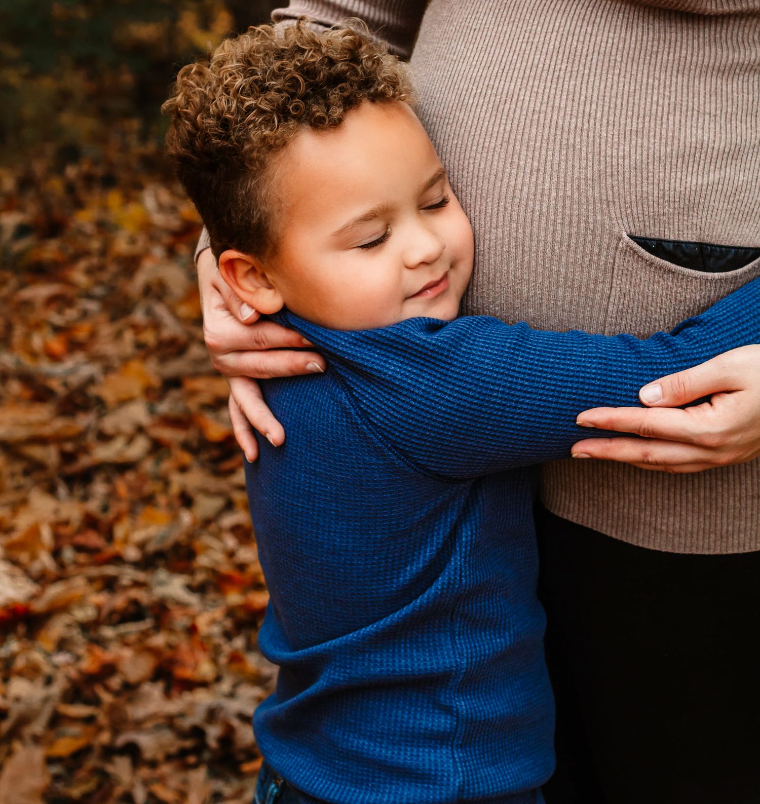 A young boy in a blue sweater hugs a pregnant woman's belly outdoors among autumn leaves.