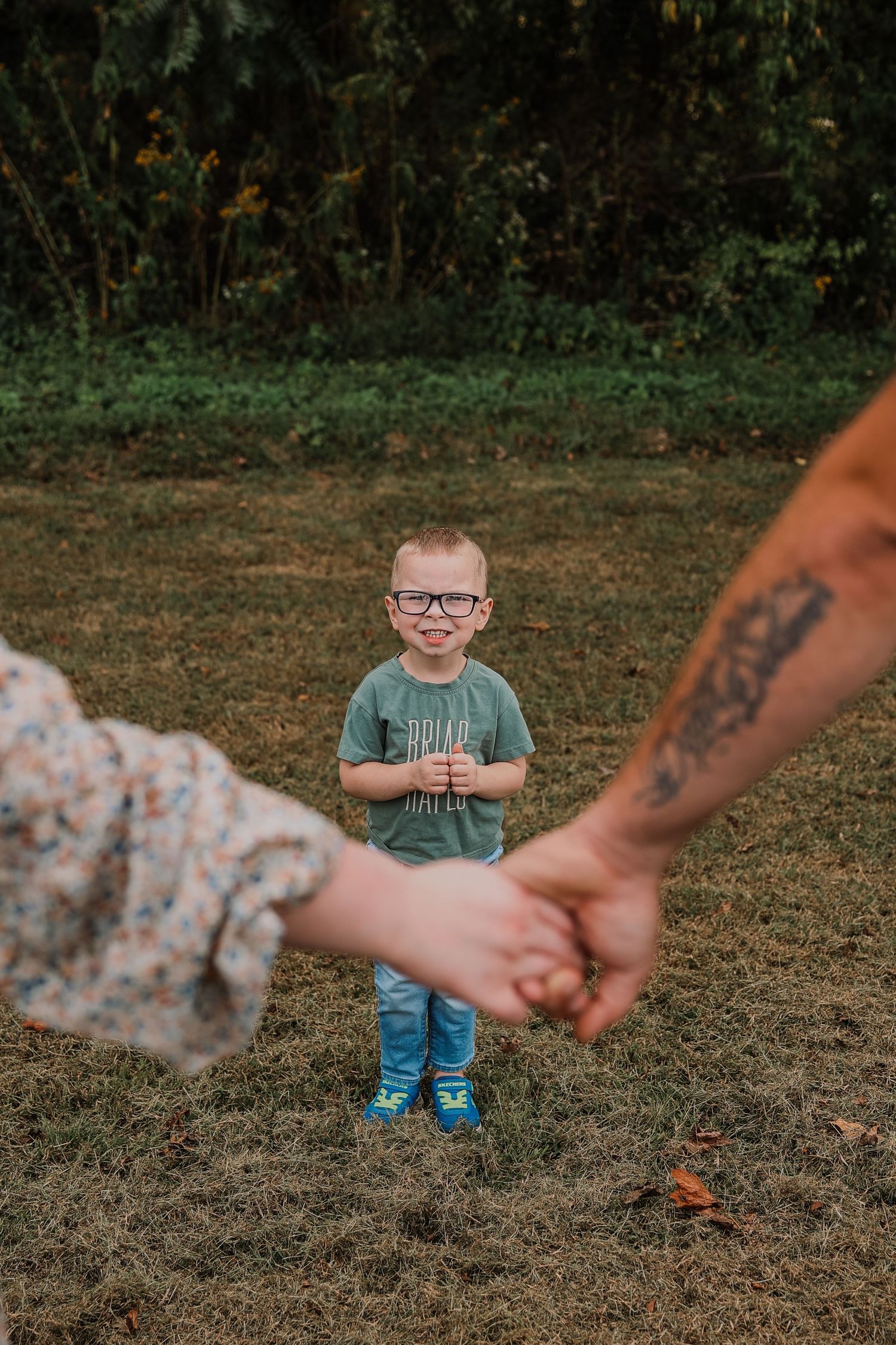 A young boy wearing glasses smiles in a grassy yard while two adults hold hands in the foreground.