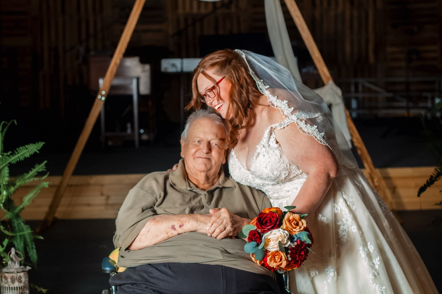Bride in white lace gown holds colorful bouquet beside elderly man in wheelchair inside rustic barn venue.