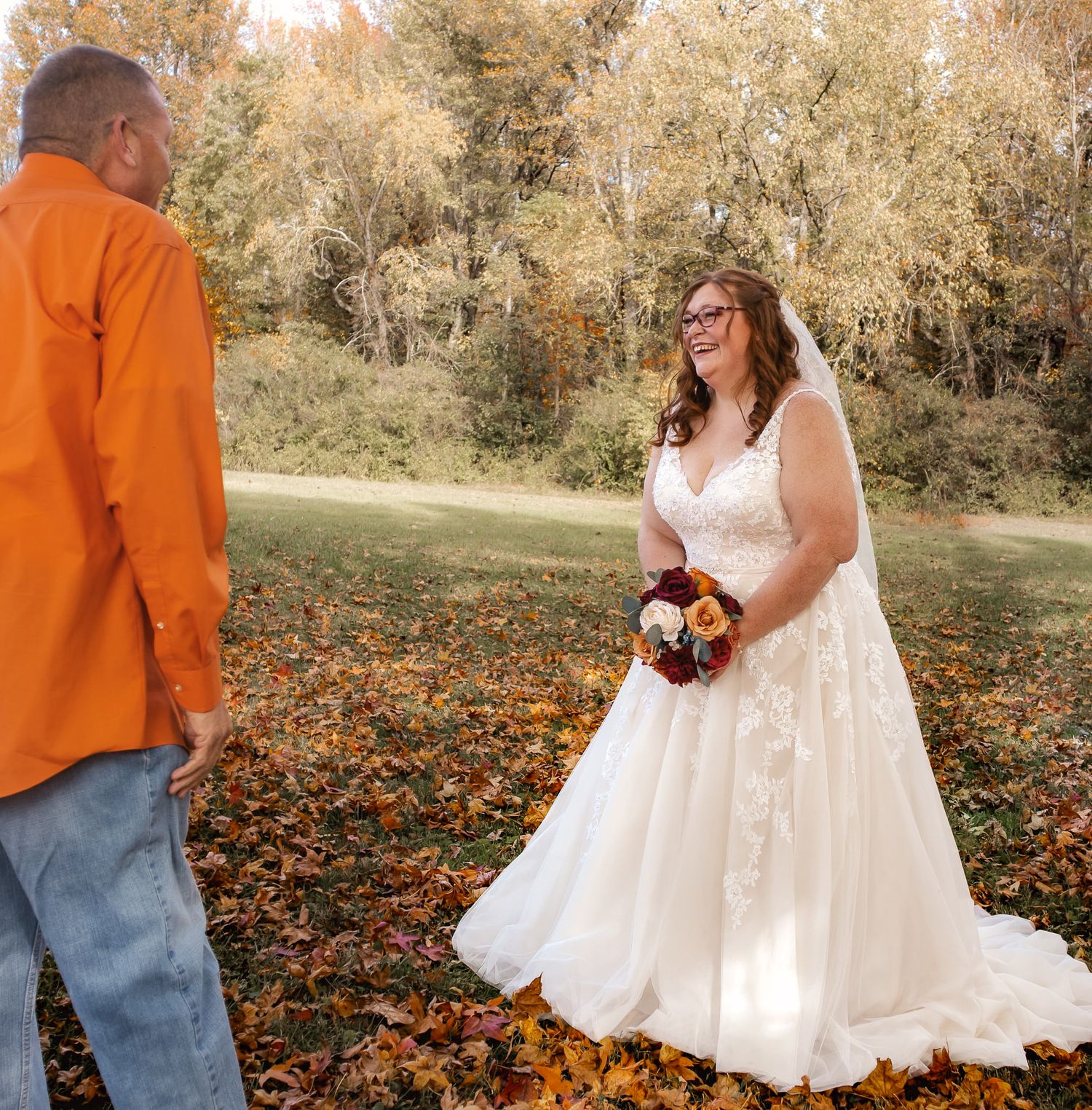 Bride in white lace gown holds bouquet facing groom in orange shirt amid autumn leaves at outdoor fall wedding.