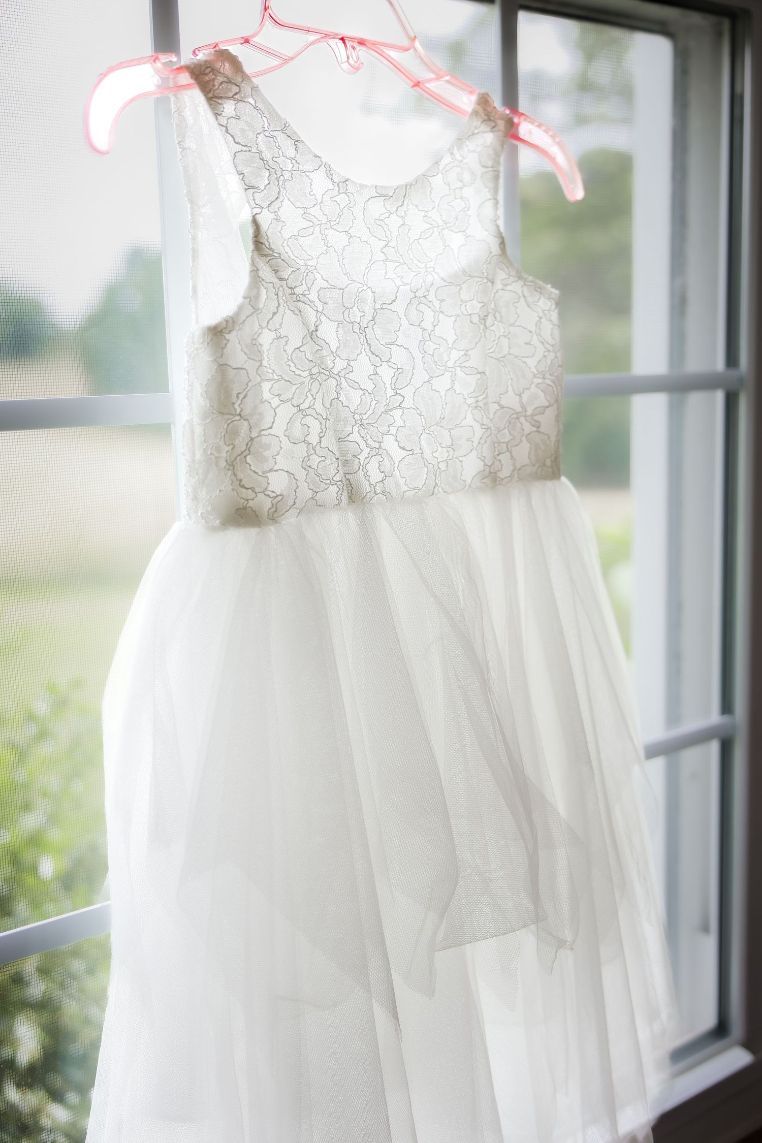 White lace and tulle flower girl dress hanging on a pink hanger in front of a window.
