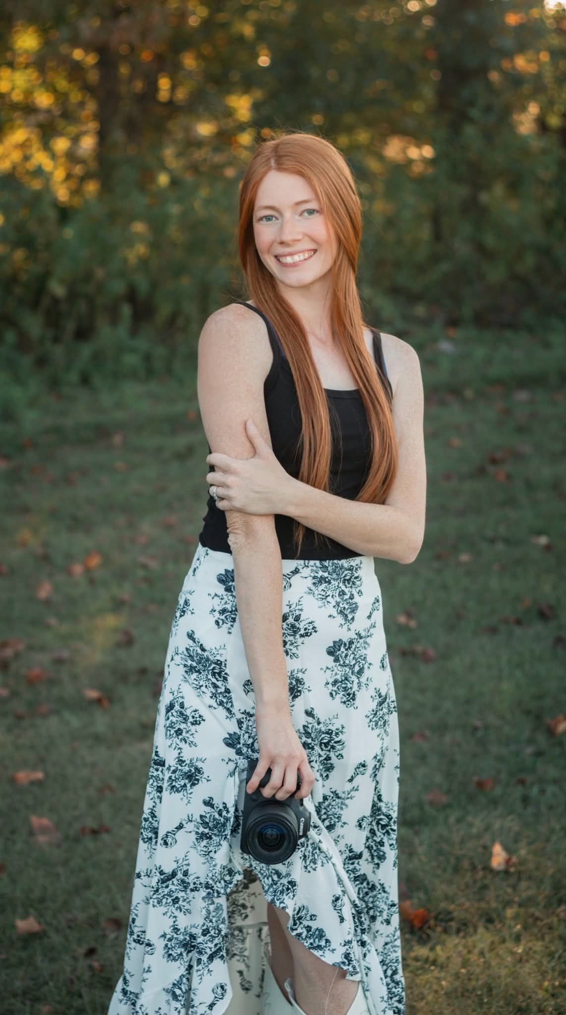 Cheerful redhead photographer in floral skirt posing with DSLR camera in green park.
