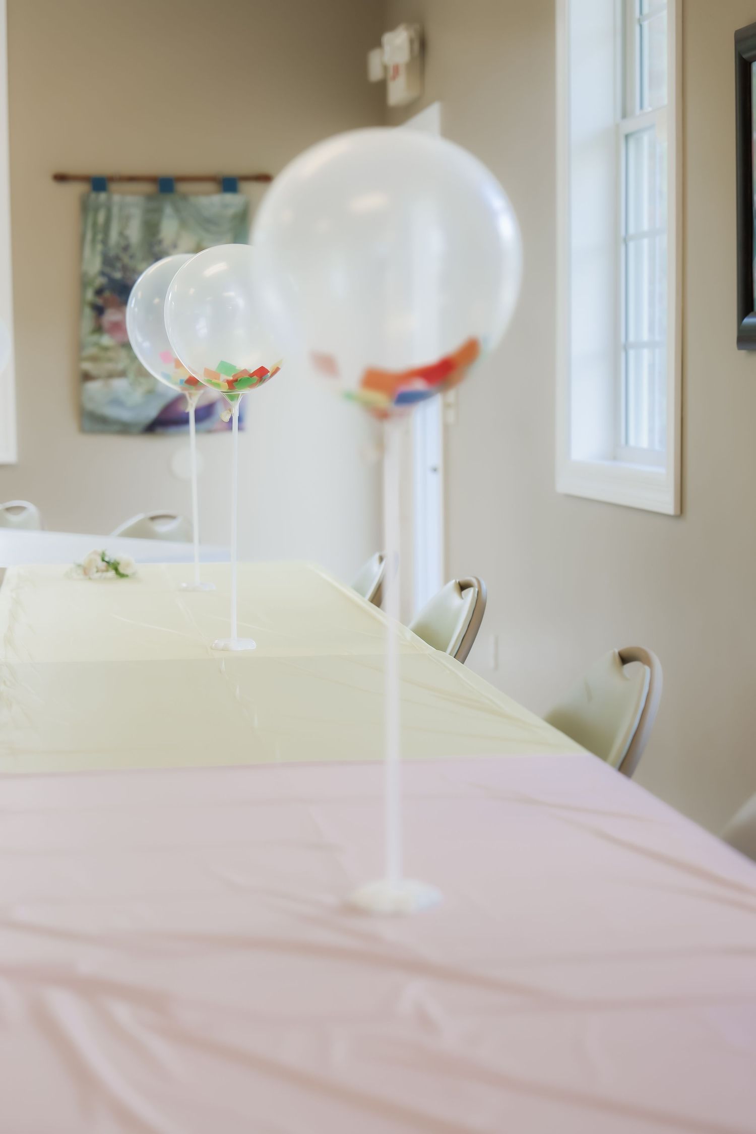 Clear balloons with colorful confetti on white stands decorate tables with pastel pink and yellow tablecloths.