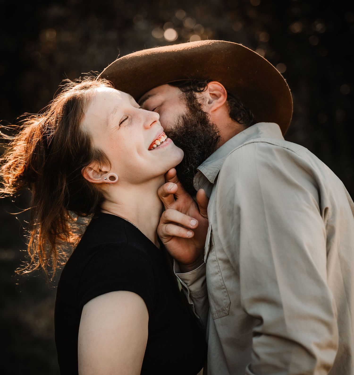 A joyful couple shares a tender moment, man in brown hat kissing smiling woman's cheek outdoors.
