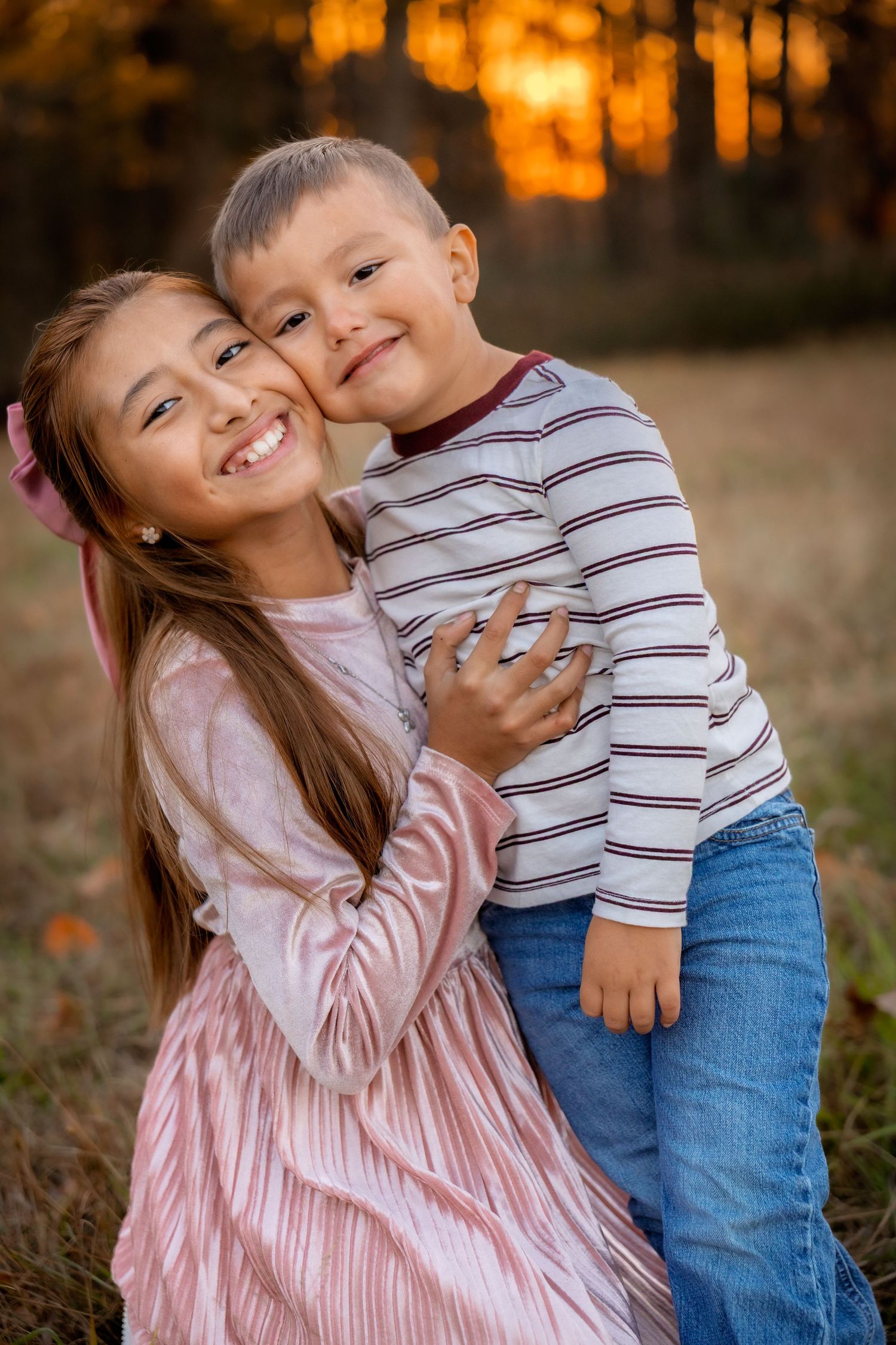 Two smiling children hugging outdoors at sunset, girl in pink dress embracing boy in striped shirt and jeans.