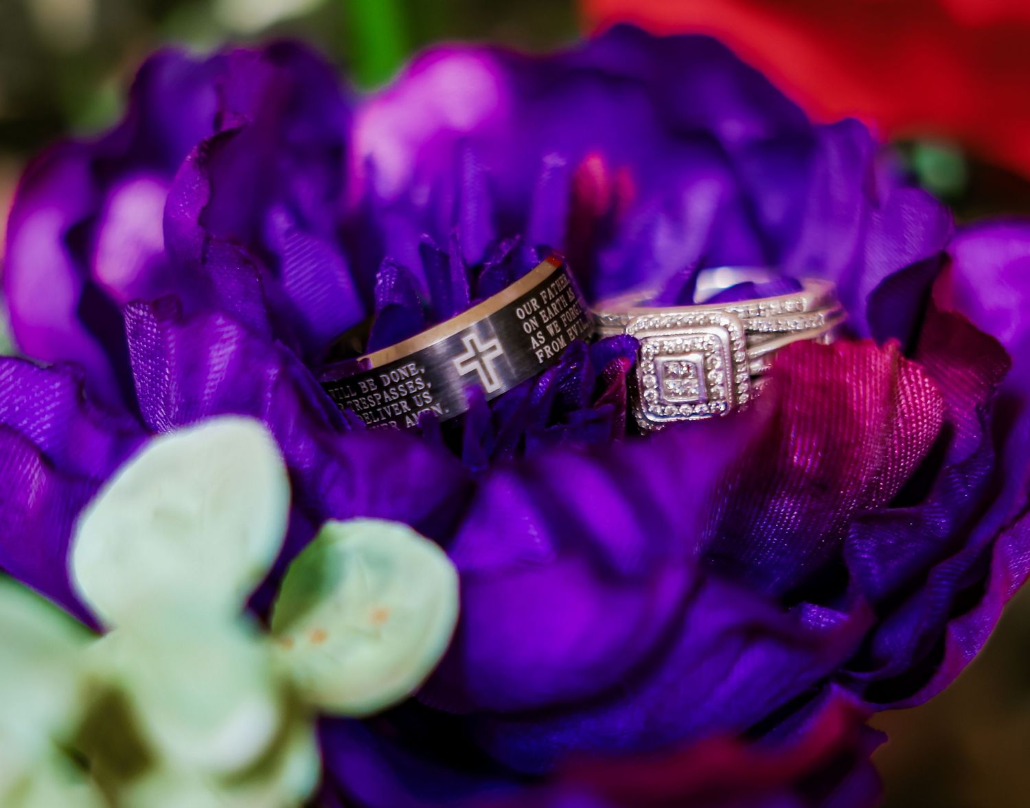 Wedding rings with cross engraving resting on vibrant purple and red flowers in a bridal bouquet arrangement.