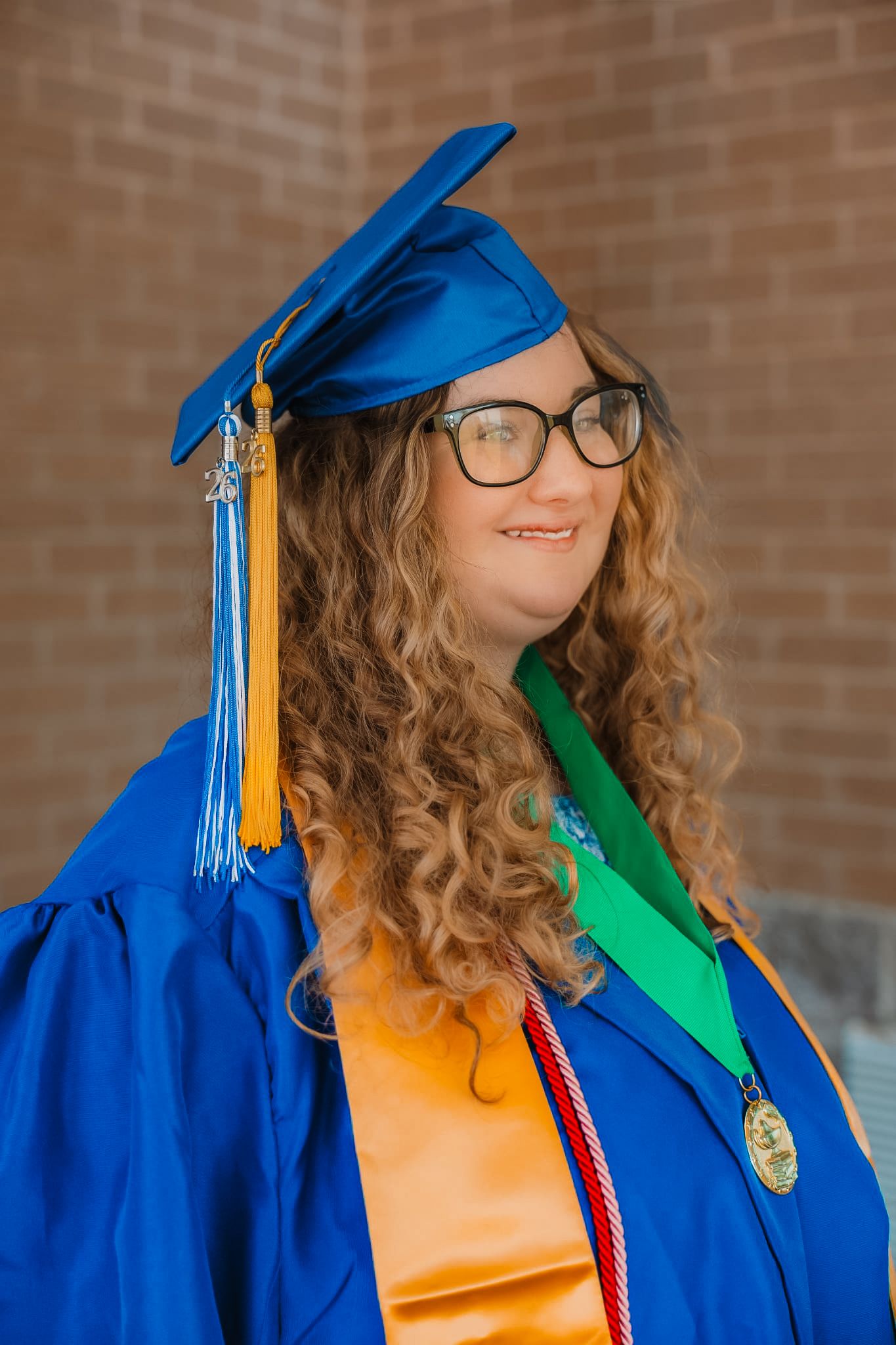 Smiling graduate in blue cap and gown with gold stole and green sash poses outdoors.