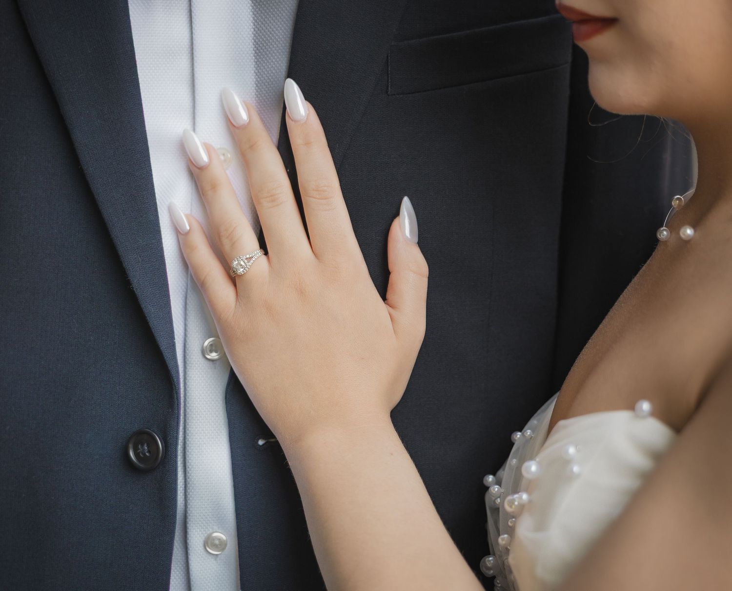 Bride with white manicured nails and diamond ring resting hand on grooms dark suit.