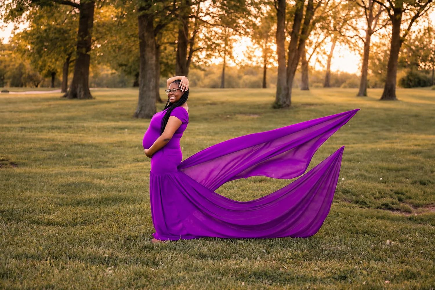 Pregnant woman in flowing purple gown poses gracefully at golden hour in a lush green park with trees.