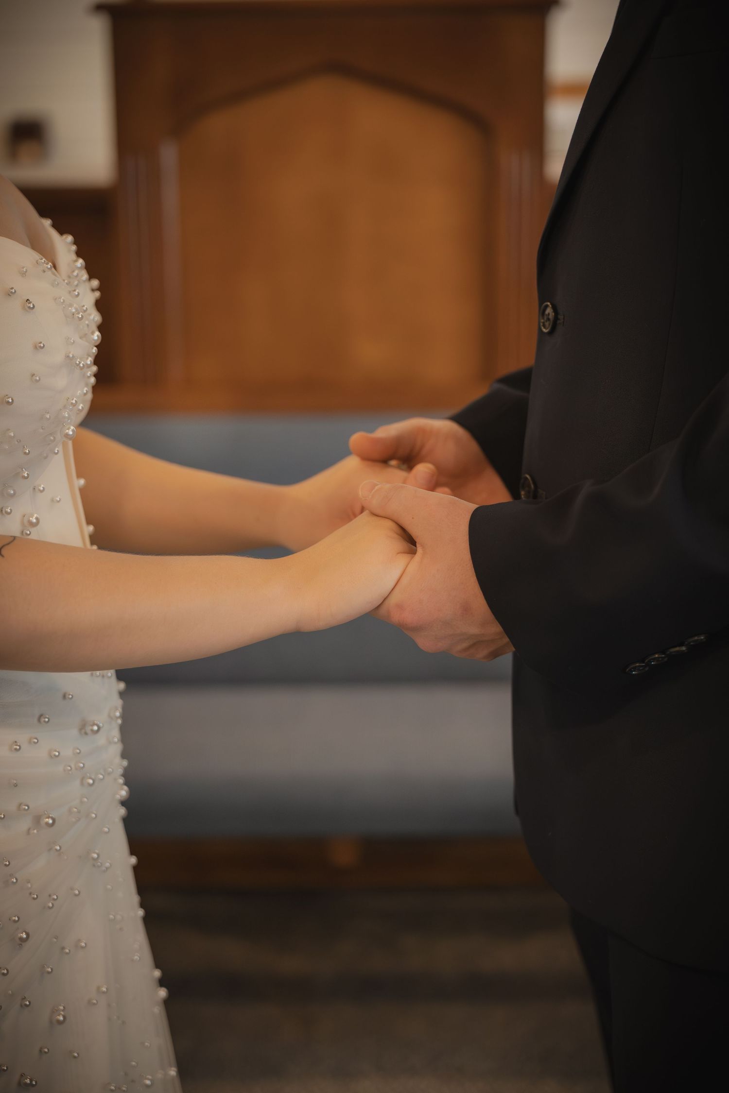 Bride and groom holding hands during wedding ceremony at church altar, bride wearing pearl-embellished white gown.