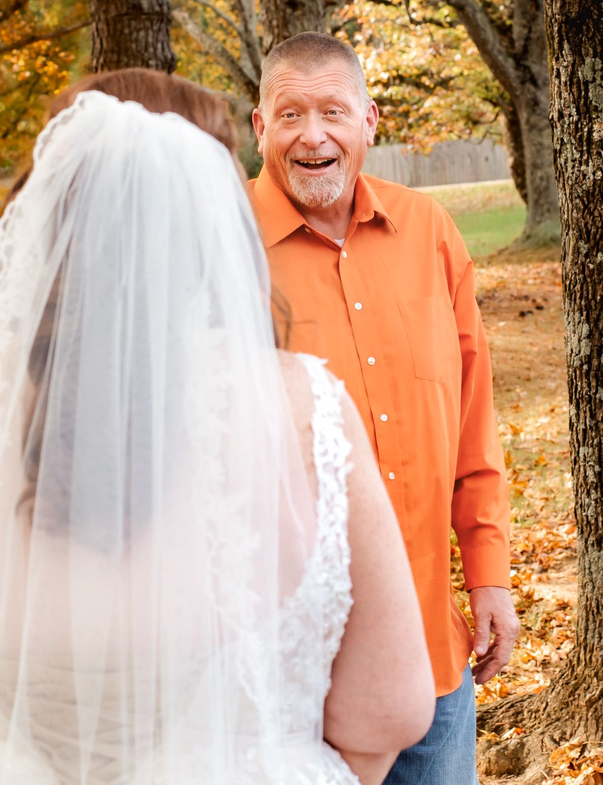 A man in an orange shirt reacts with joy seeing a bride in a white lace gown and veil outdoors in autumn.