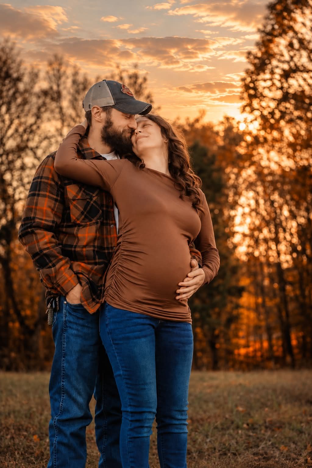 Couple embracing at sunset during maternity photo shoot in autumn field, woman in brown top cradling baby bump.
