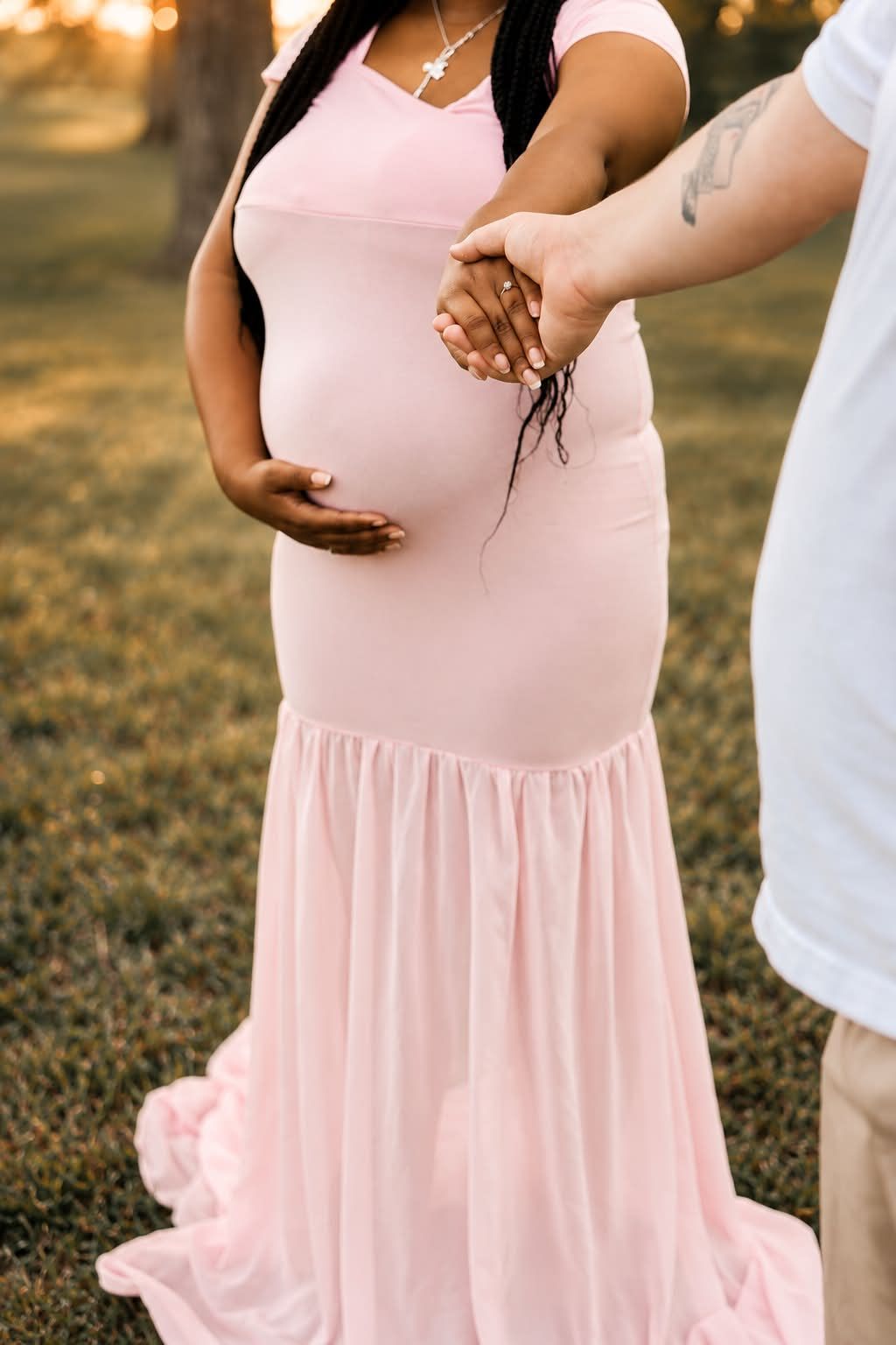 Pregnant woman in pink maternity gown holds hands with partner outdoors at golden hour.