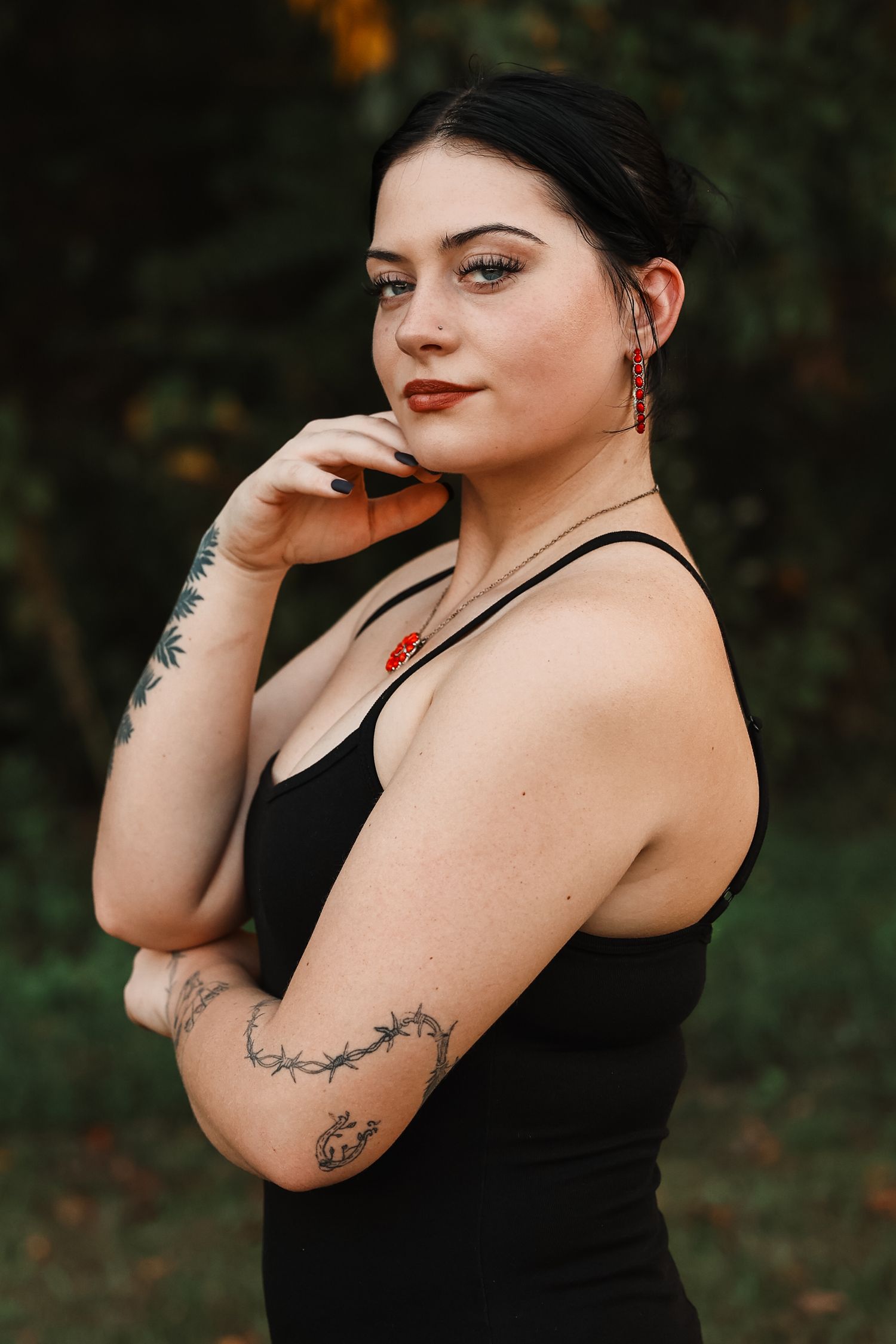 Young woman with tattoos in black dress poses outdoors with hand near face, wearing red earrings.