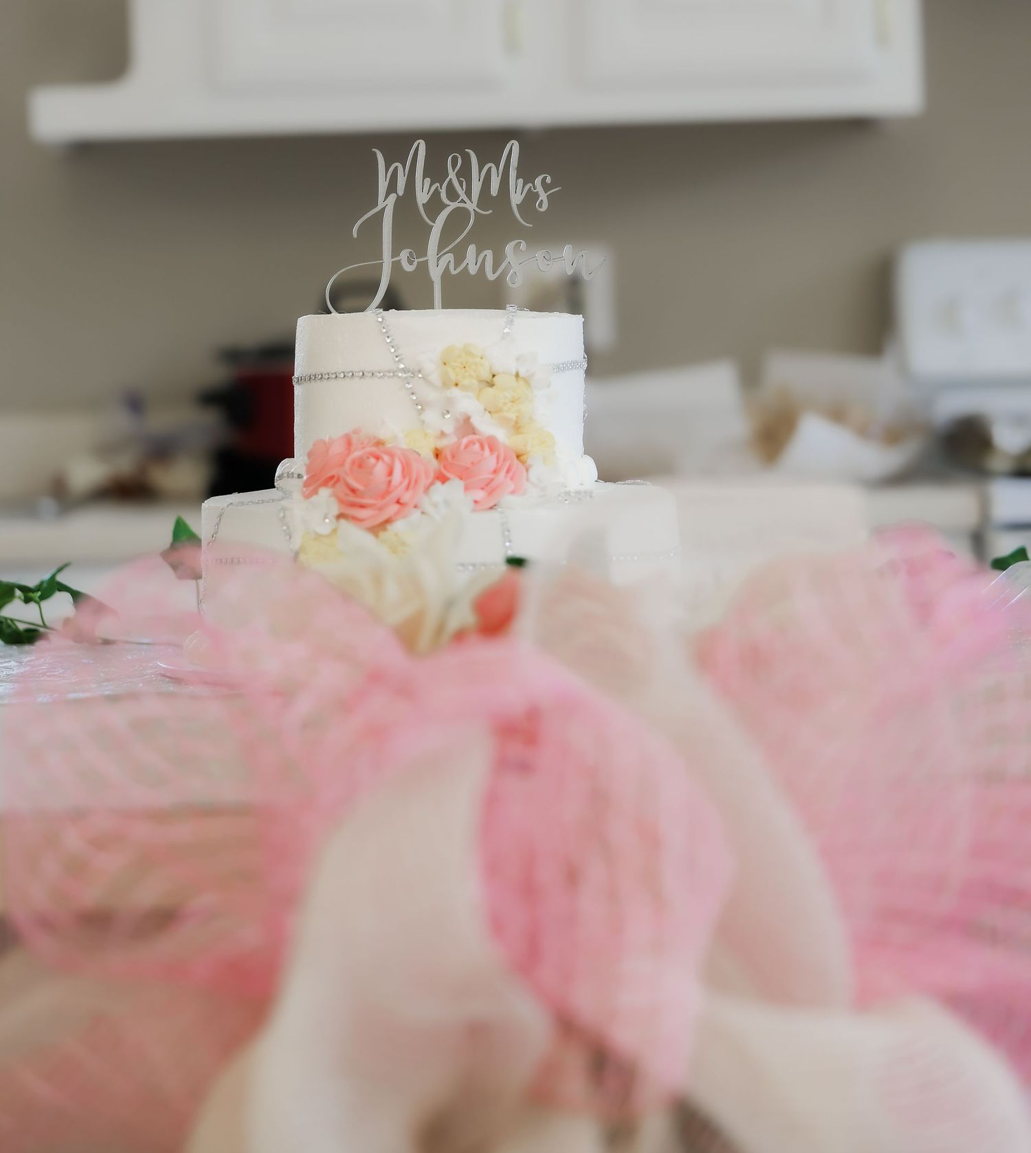 Two-tier white wedding cake with pink roses and Mr & Mrs Johnson topper, surrounded by pink tulle decorations.