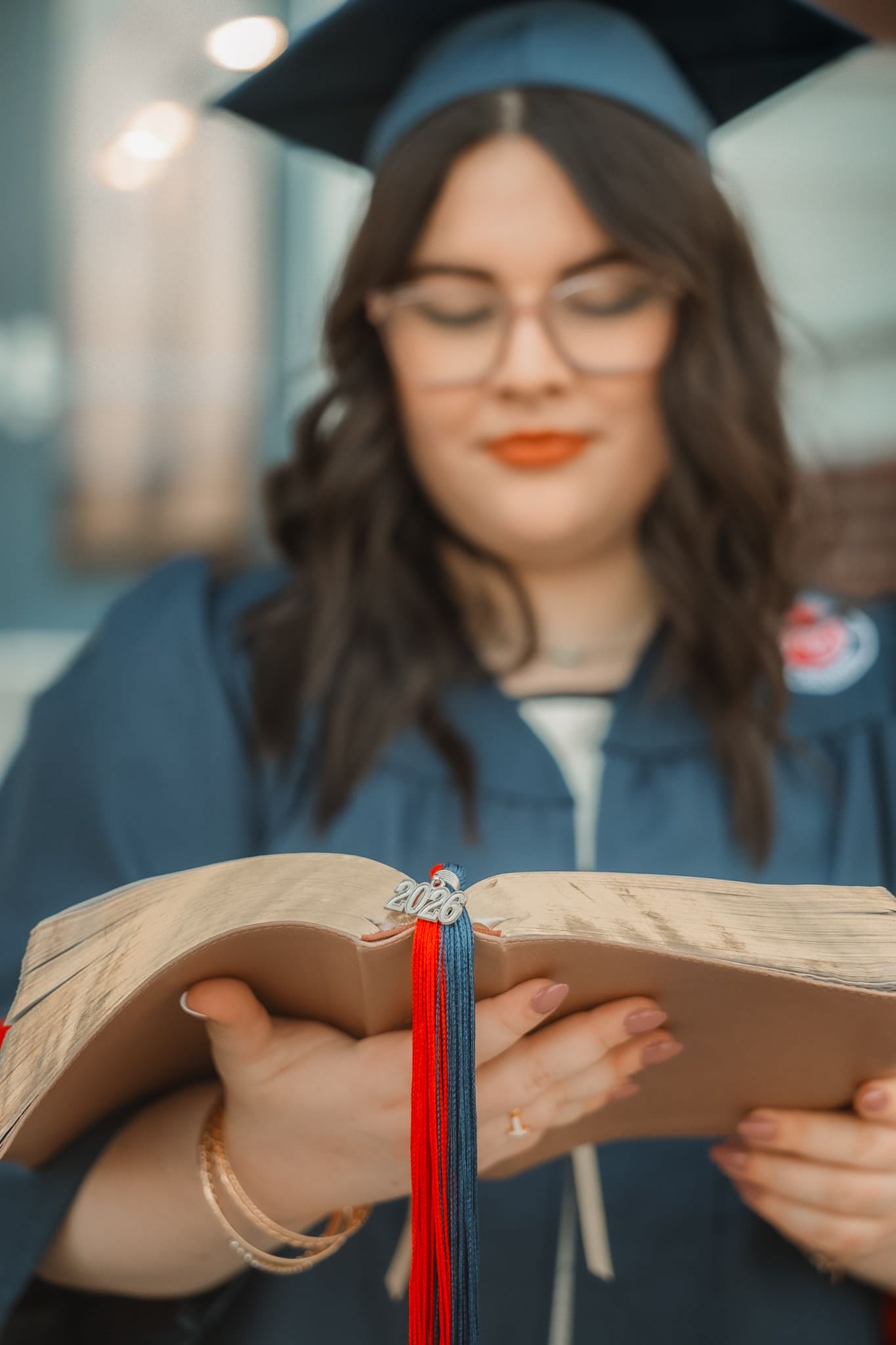 A graduate in blue cap and gown reads an open book with red and blue tassel, wearing glasses and orange lipstick.