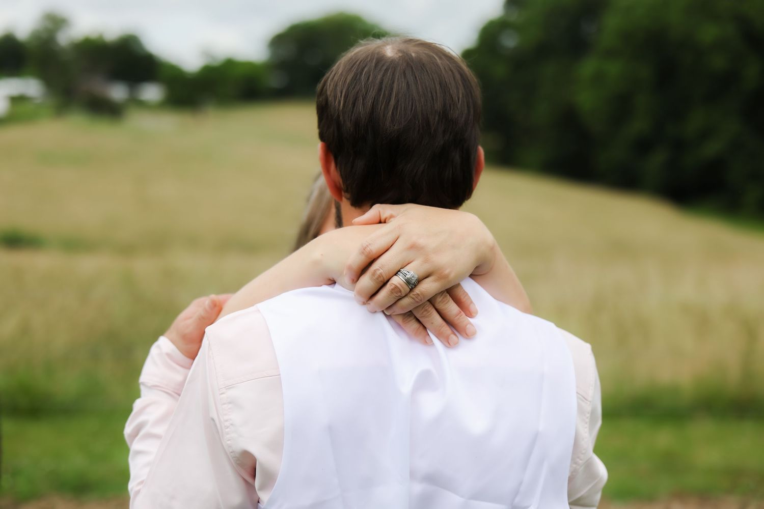 Couple embracing outdoors in a field, woman wearing an engagement ring, viewed from behind.