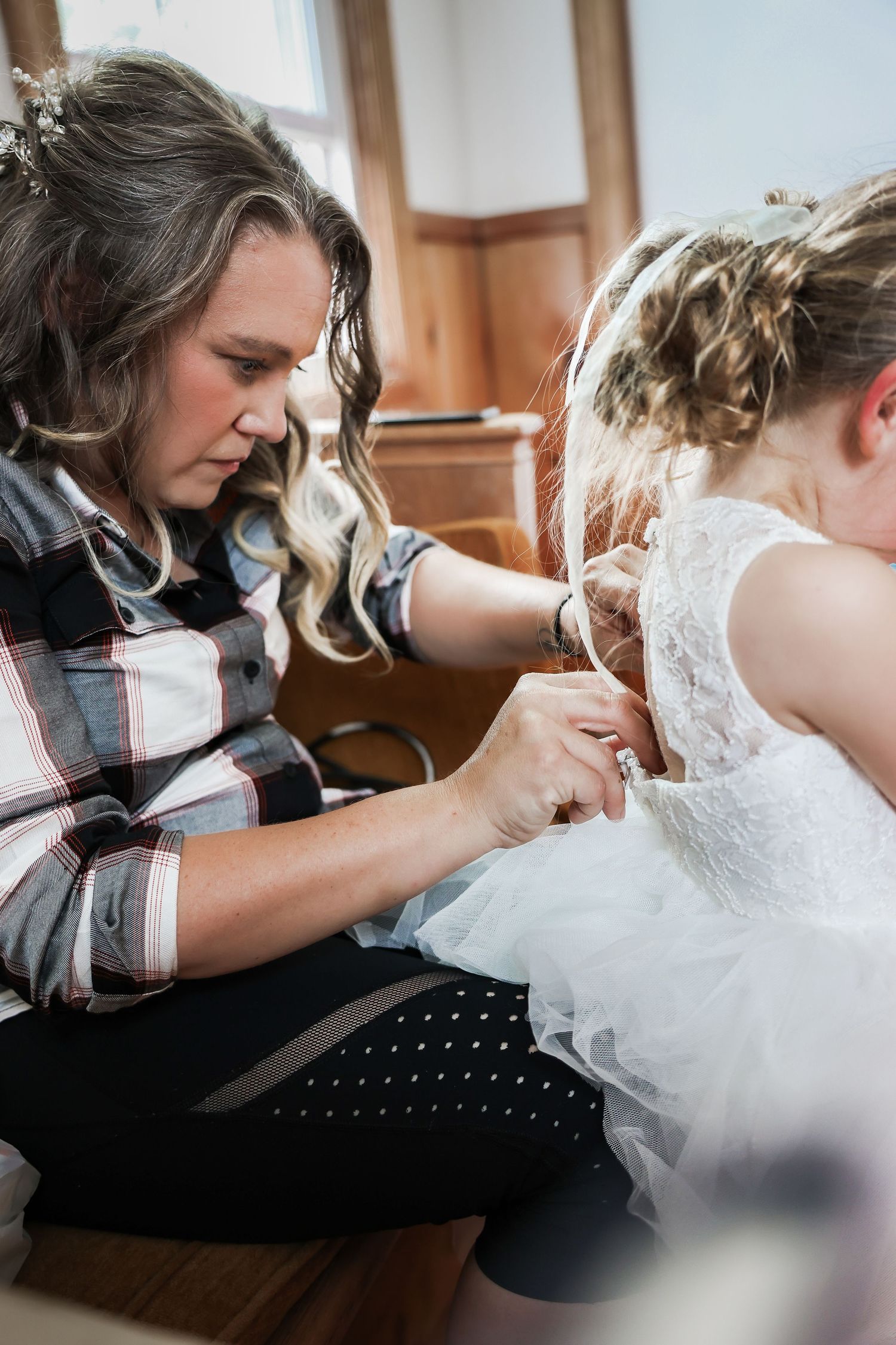 Woman in plaid shirt carefully buttoning the back of a young girl's white lace flower girl dress.