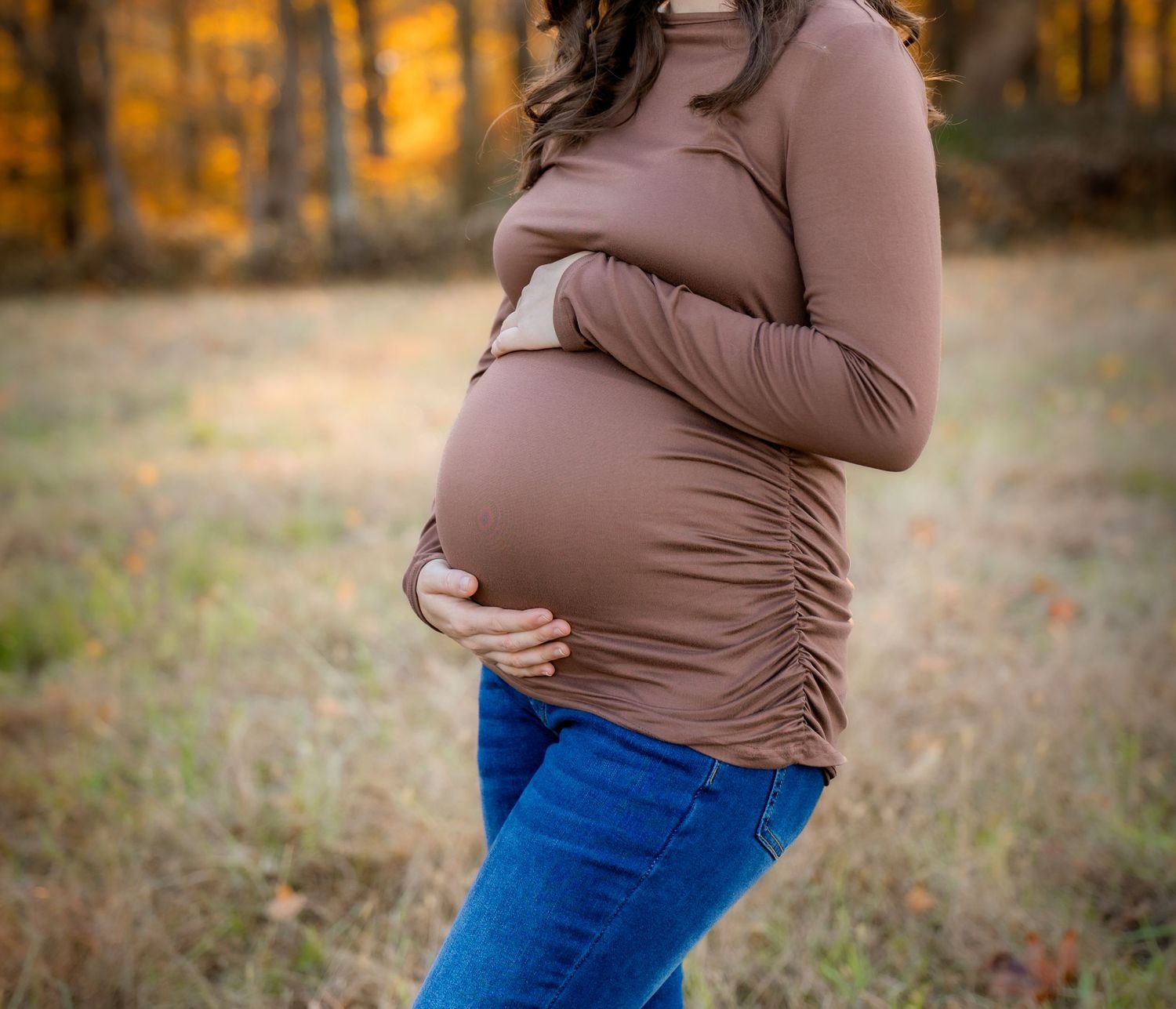 Pregnant woman in brown turtleneck and jeans cradling her baby bump outdoors in an autumn forest setting.